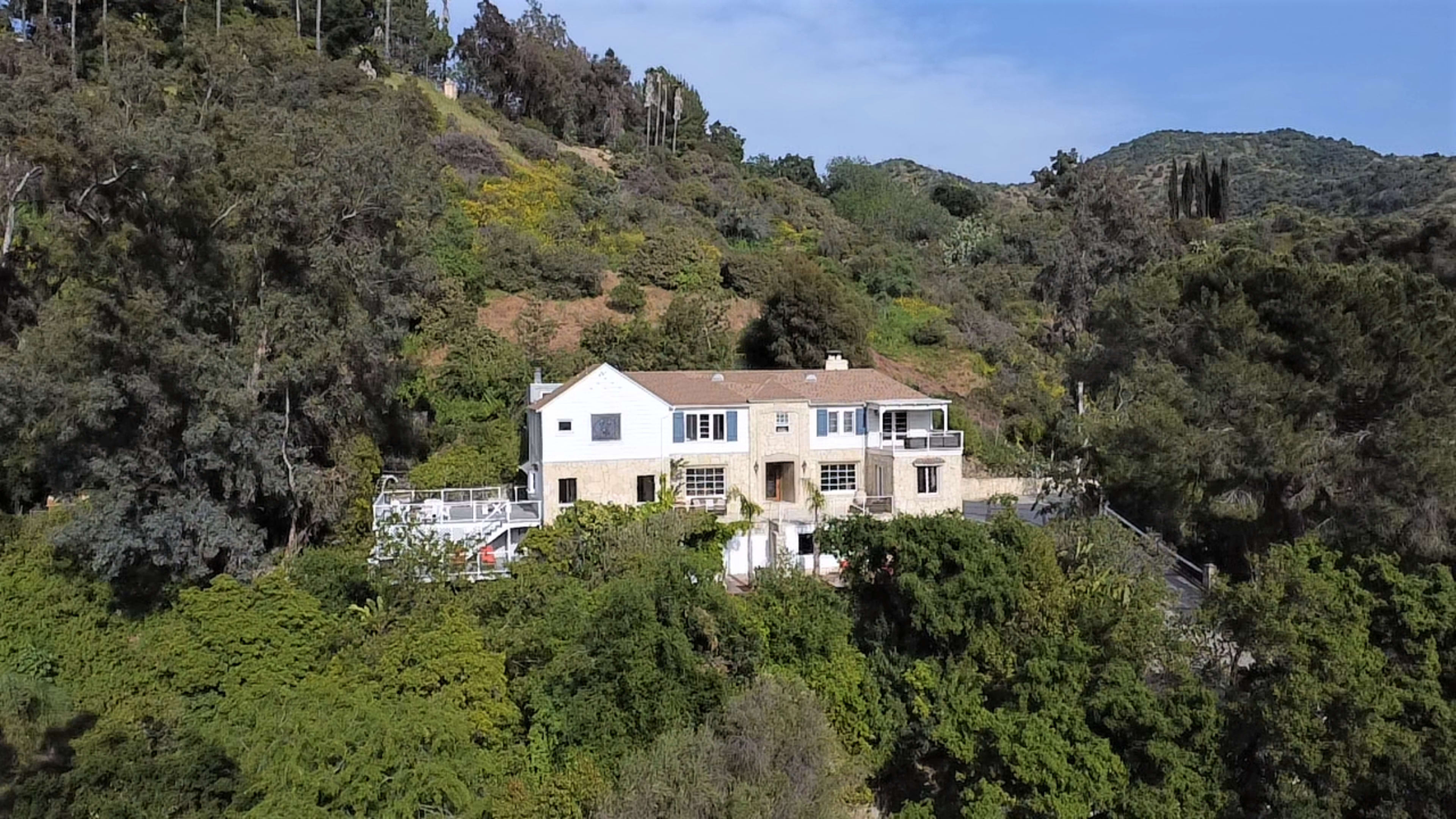 A large white house with a tan roof is surrounded by dense greenery and situated on a hillside.