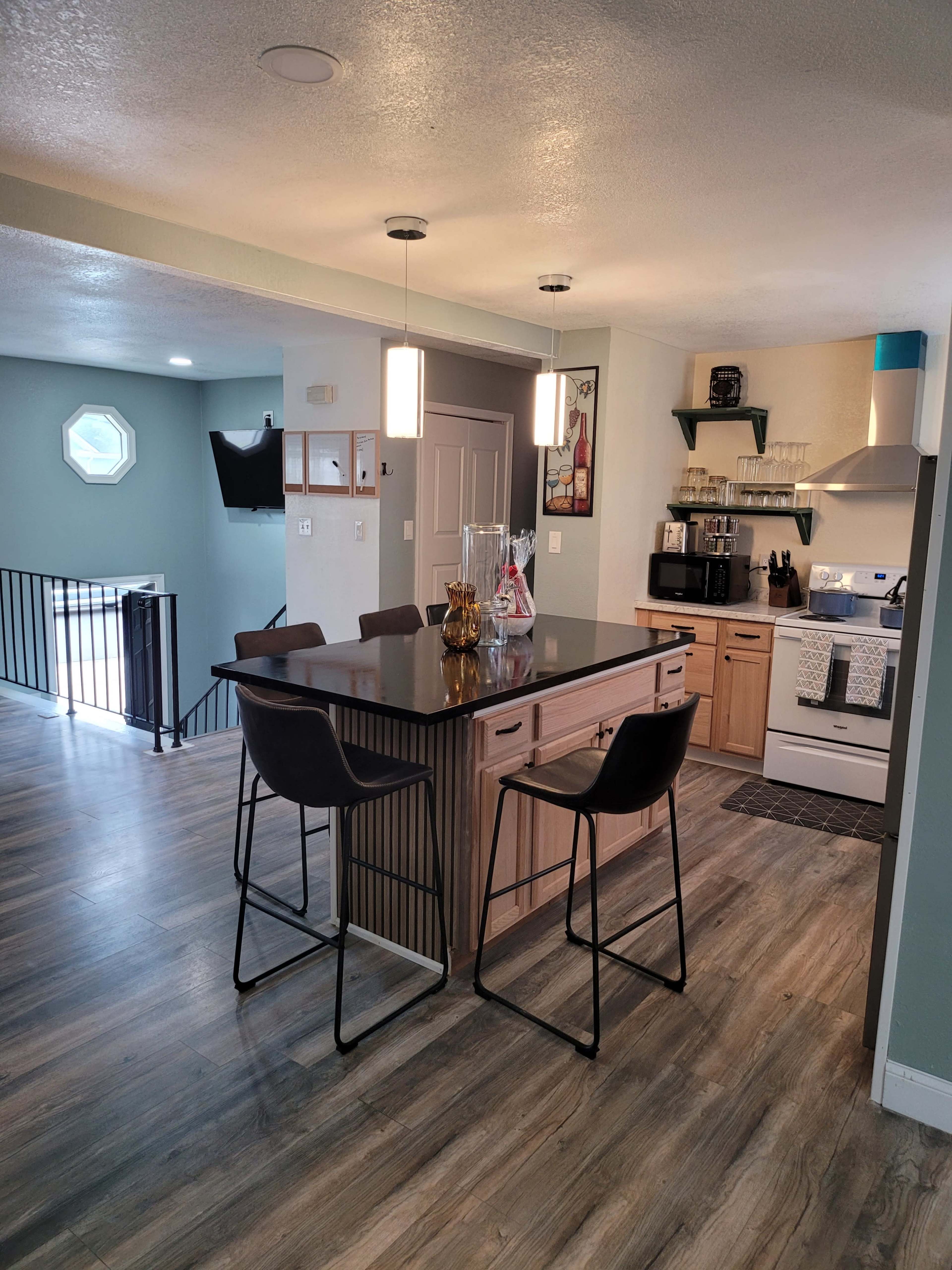 The image shows a modern kitchen with an island bar, dark countertops, and seating for two, adjacent to a dining area and a staircase in the background.