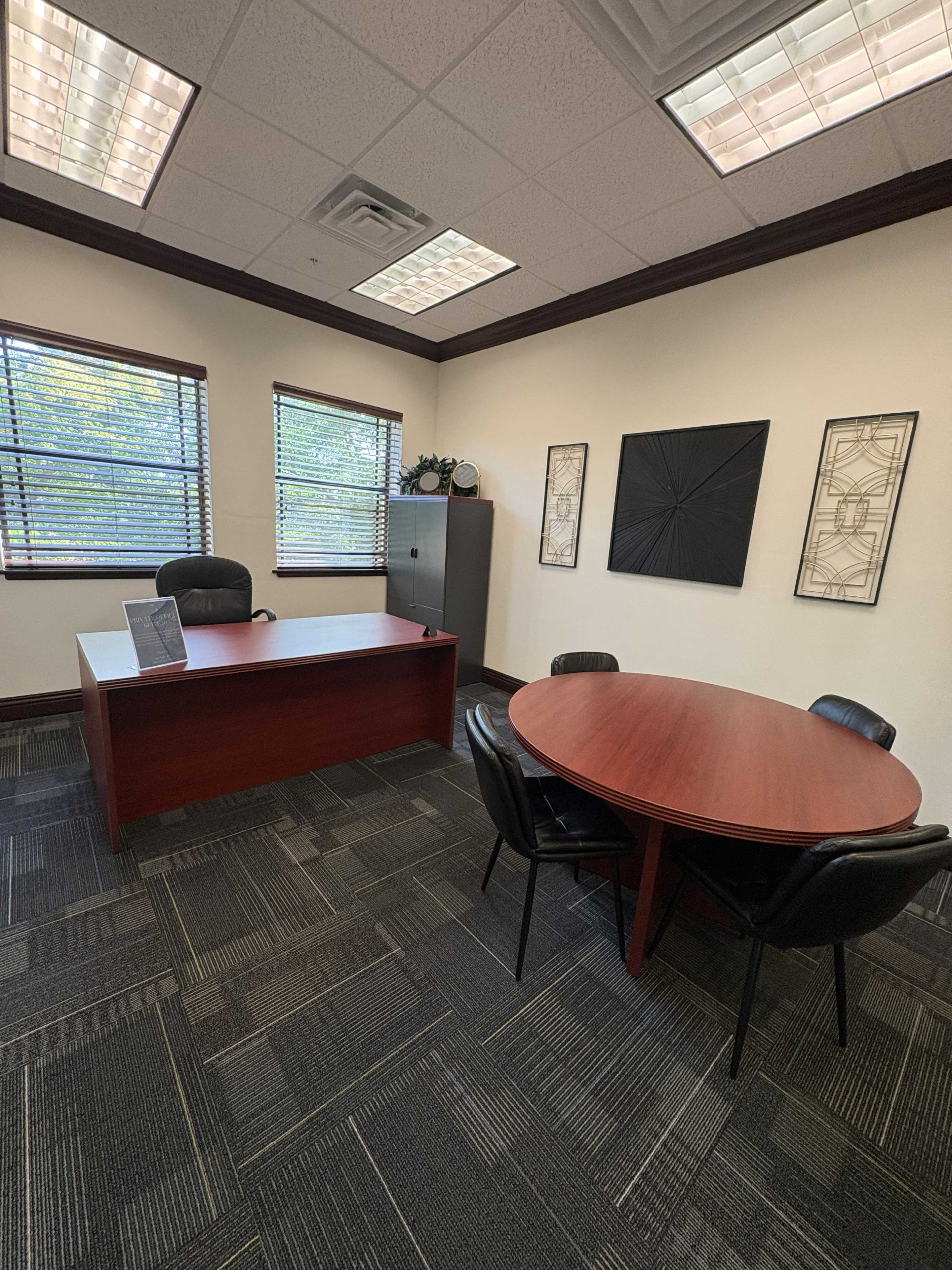 The image shows an office space featuring a wooden desk and chair, a round table with four black chairs, and two windows with blinds.