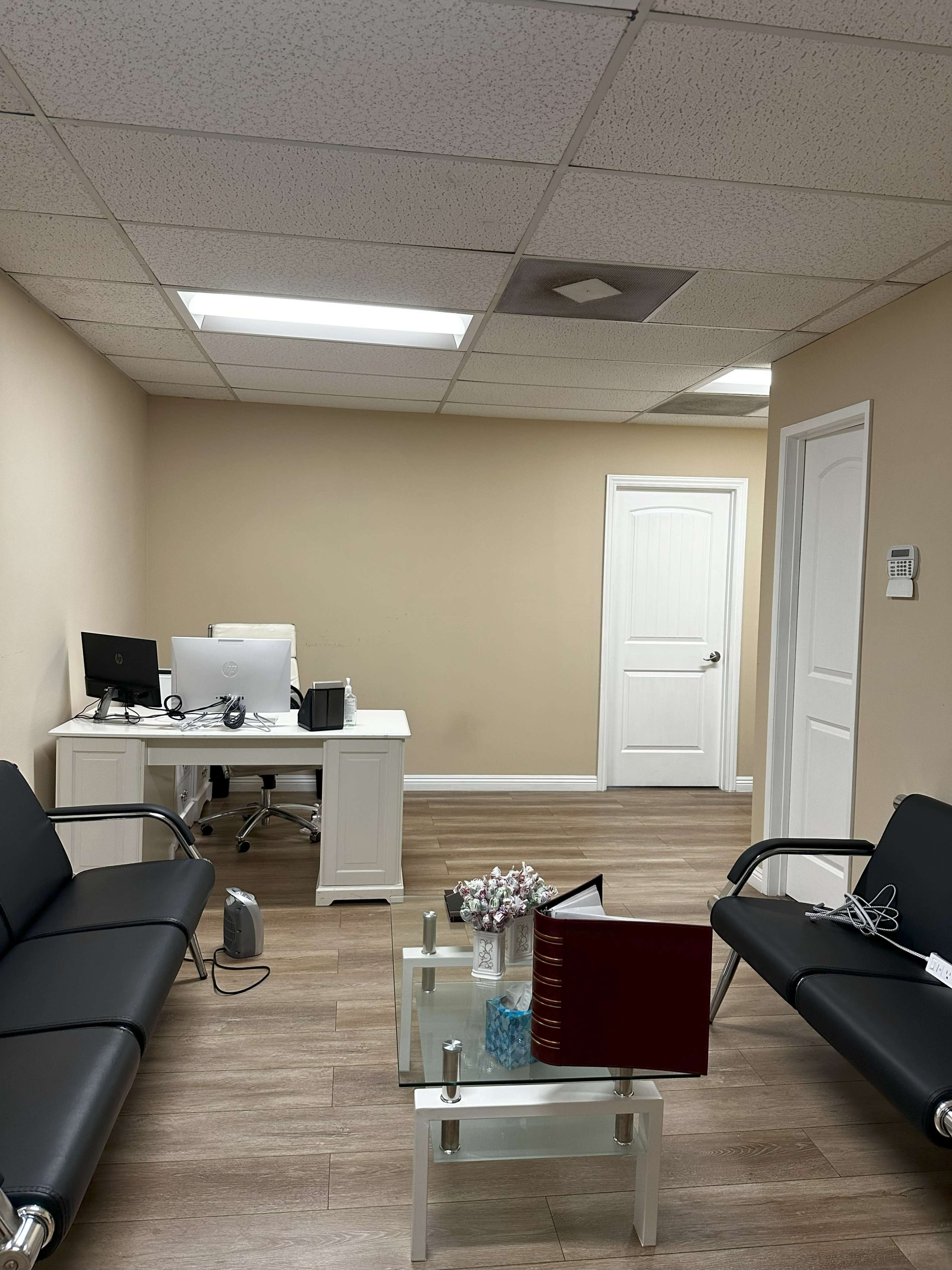 The image shows a waiting room with black leather sofas, a glass coffee table, a desk with a computer, and a door leading to another room.
