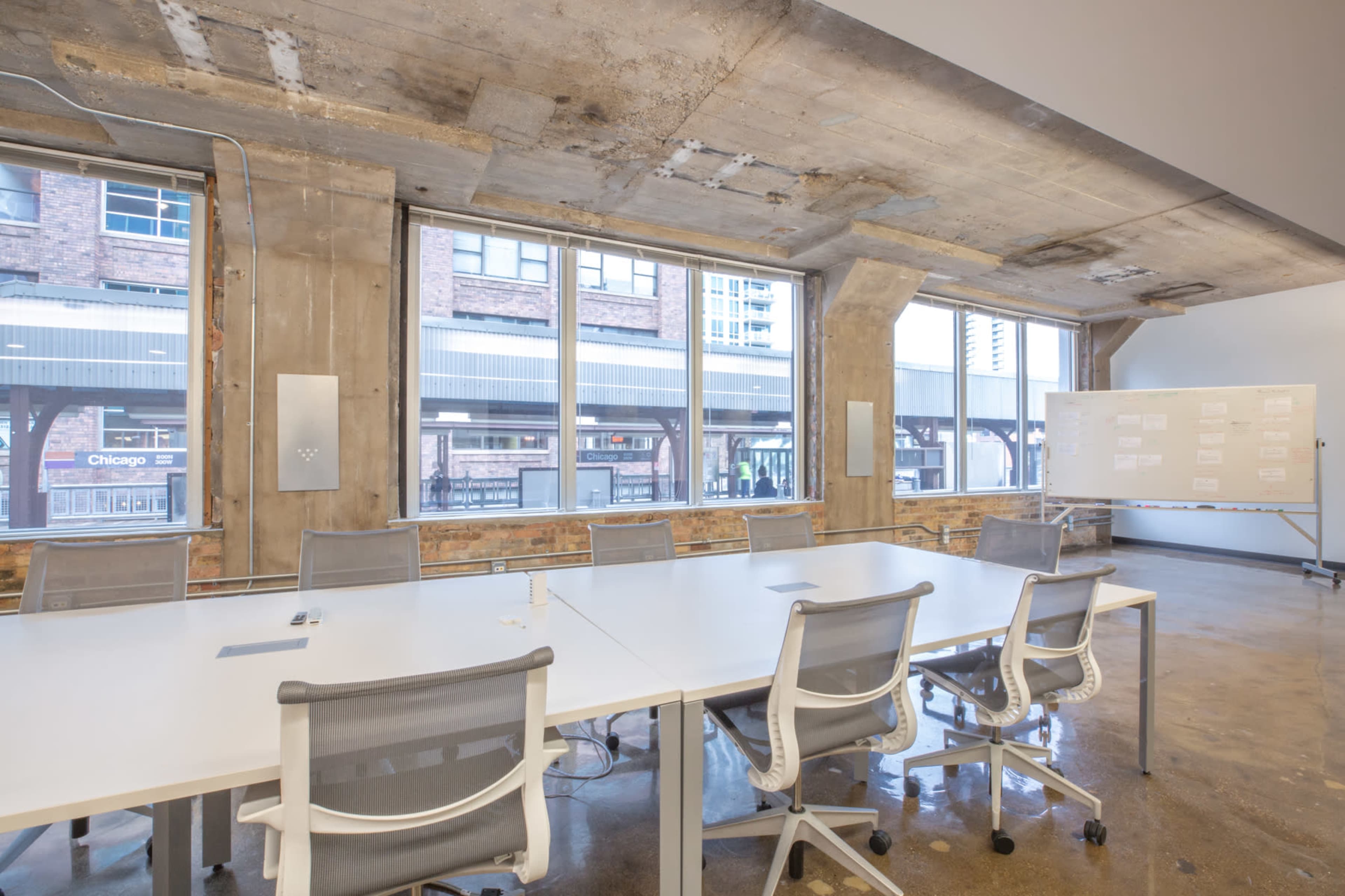 A modern conference room with large windows, a long white table, and ergonomic chairs, featuring exposed concrete walls and a whiteboard.