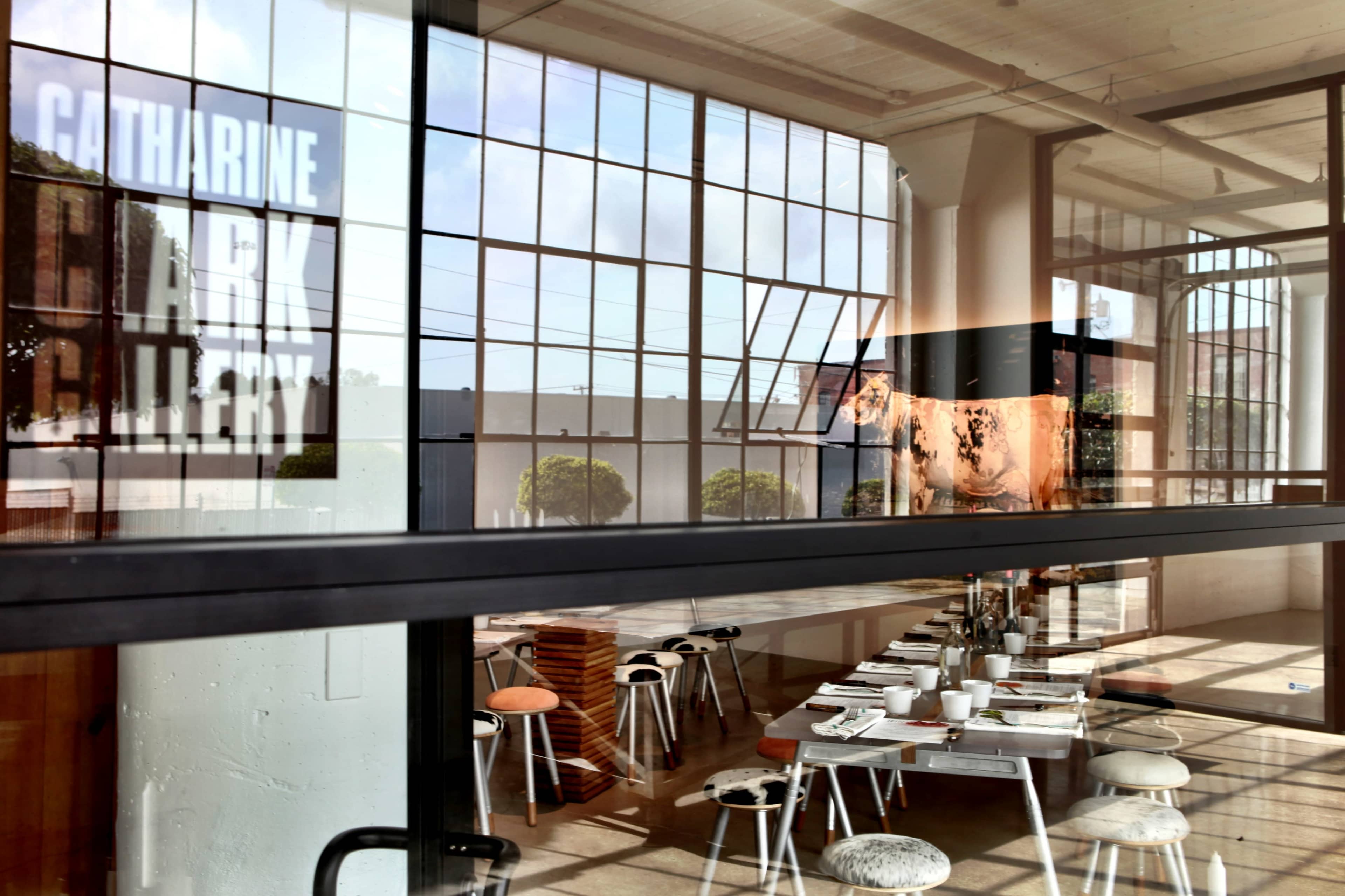 The image shows a modern art gallery with large windows and a dining area visible through the glass, featuring a long table set with plates and chairs.
