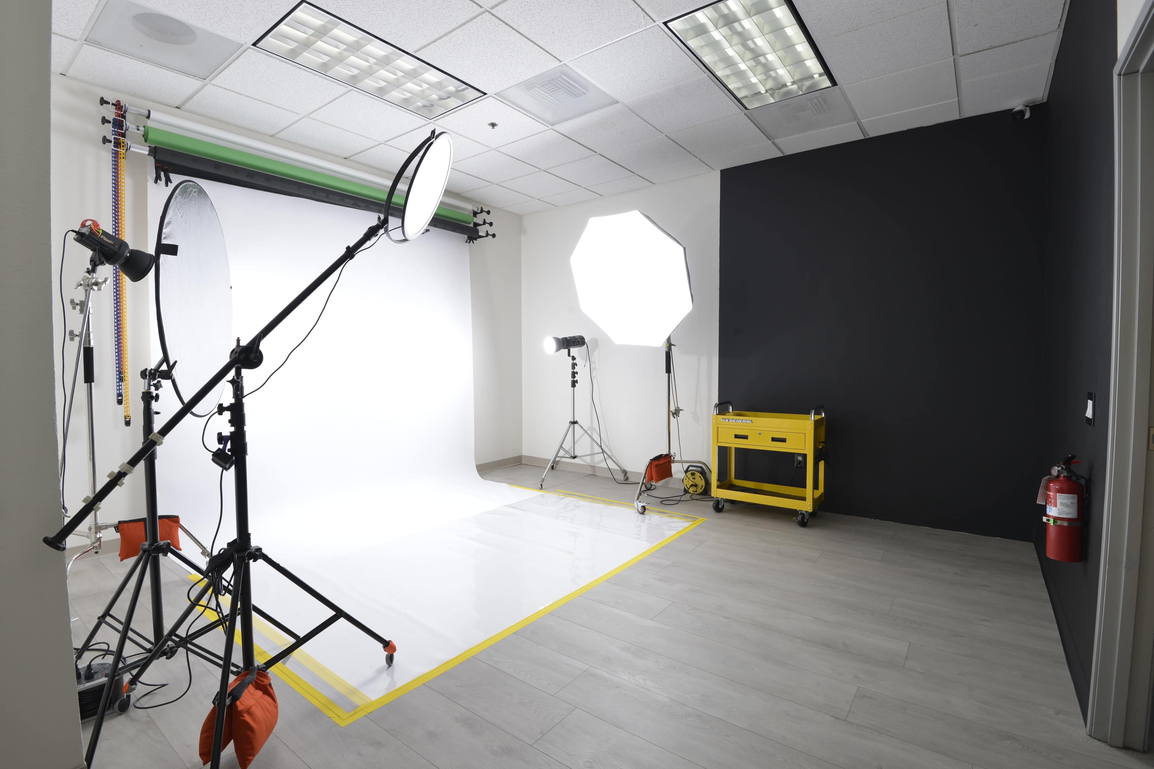 The image shows a photo studio setup with a white backdrop, several lighting equipment, and a black wall on one side.