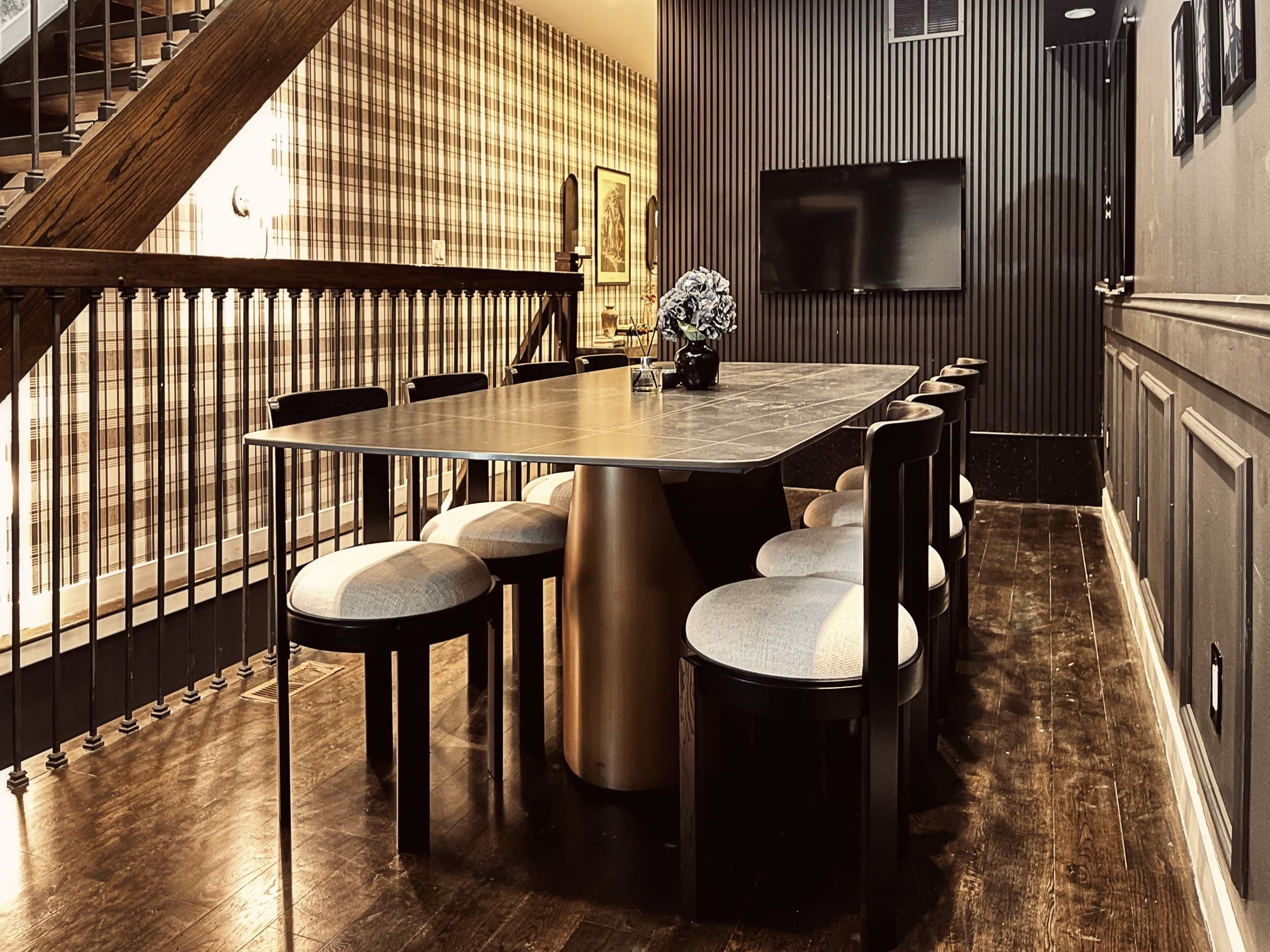 A modern dining area features a large table surrounded by eight upholstered chairs, with a staircase and patterned wallpaper in the background.