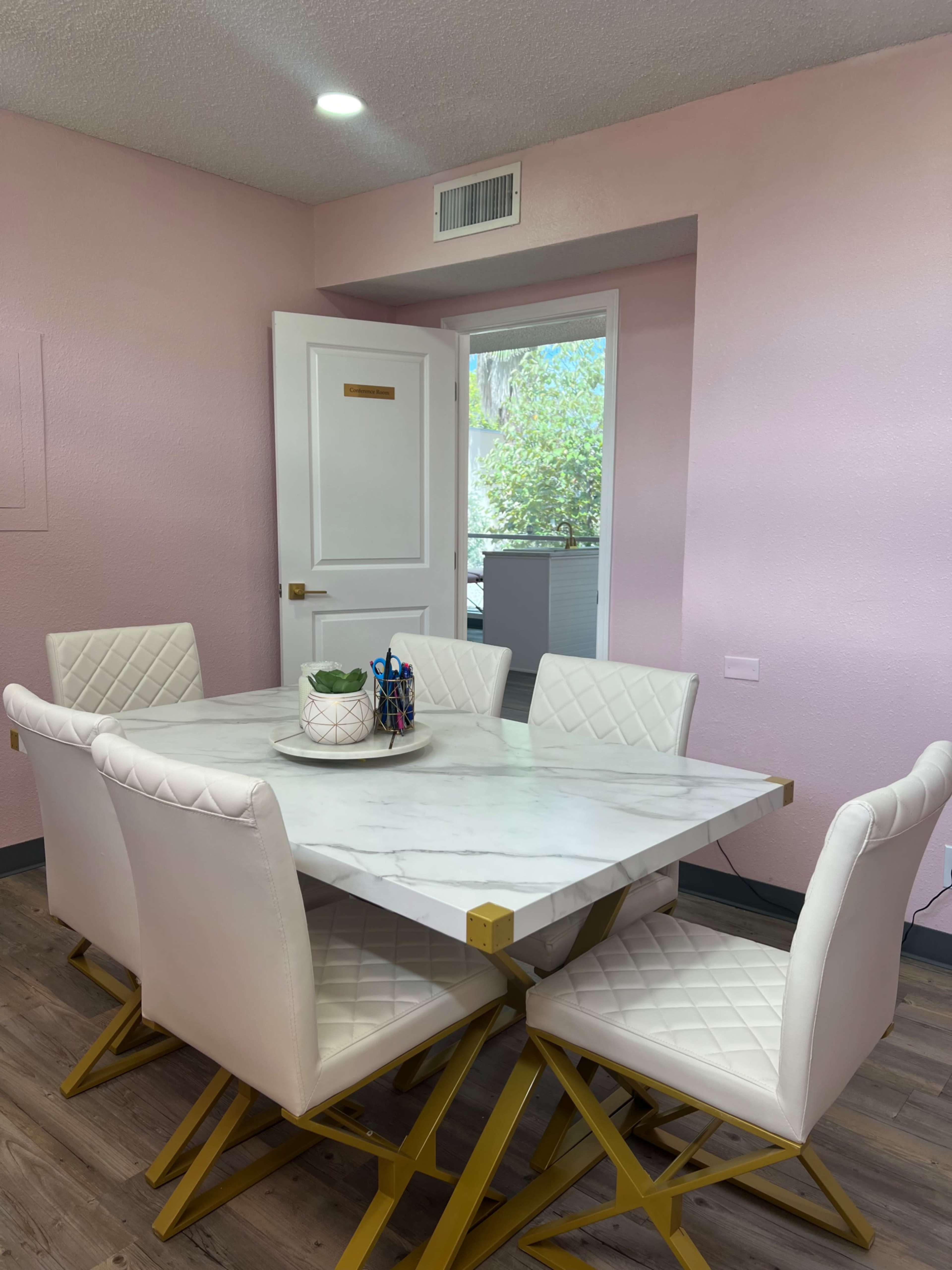 A modern dining room features a square marble table surrounded by white upholstered chairs, against a light pink wall.