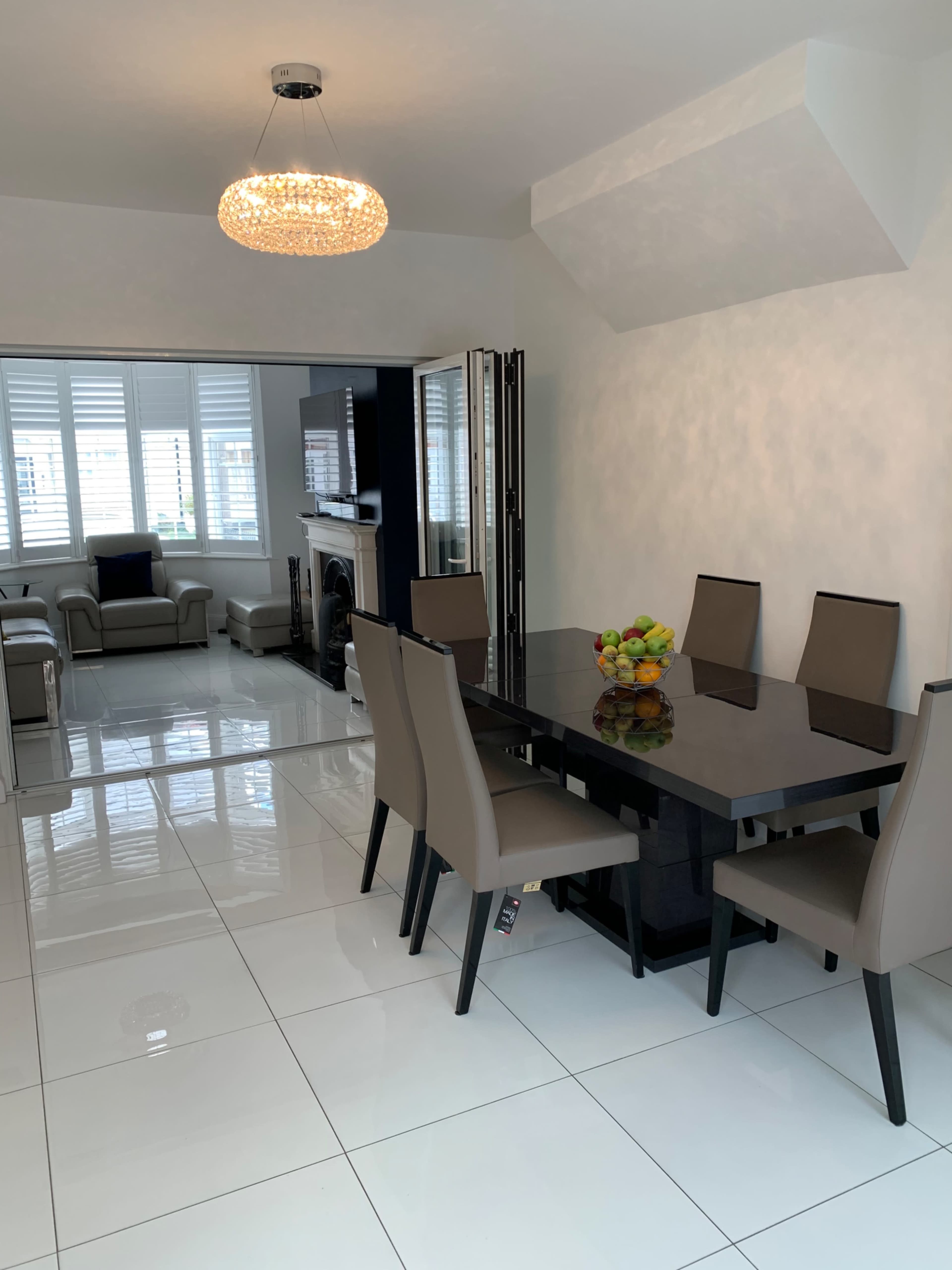 A modern dining area with a sleek black table, six beige chairs, and a bowl of fruit as a centerpiece, all situated on shiny white tiles.