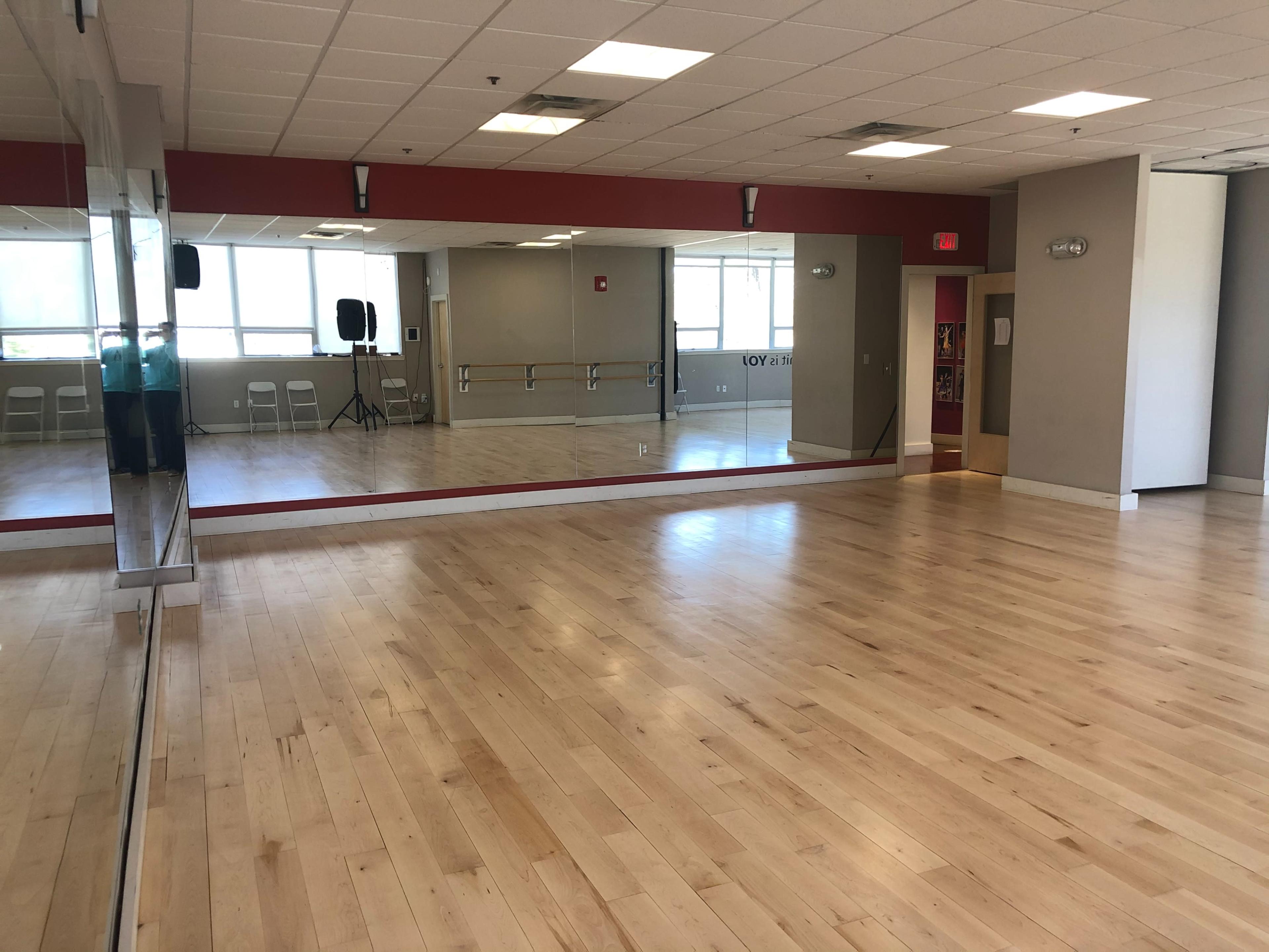 The image shows a spacious dance studio featuring wooden floors, mirrors on one wall, and a few chairs arranged in the background.