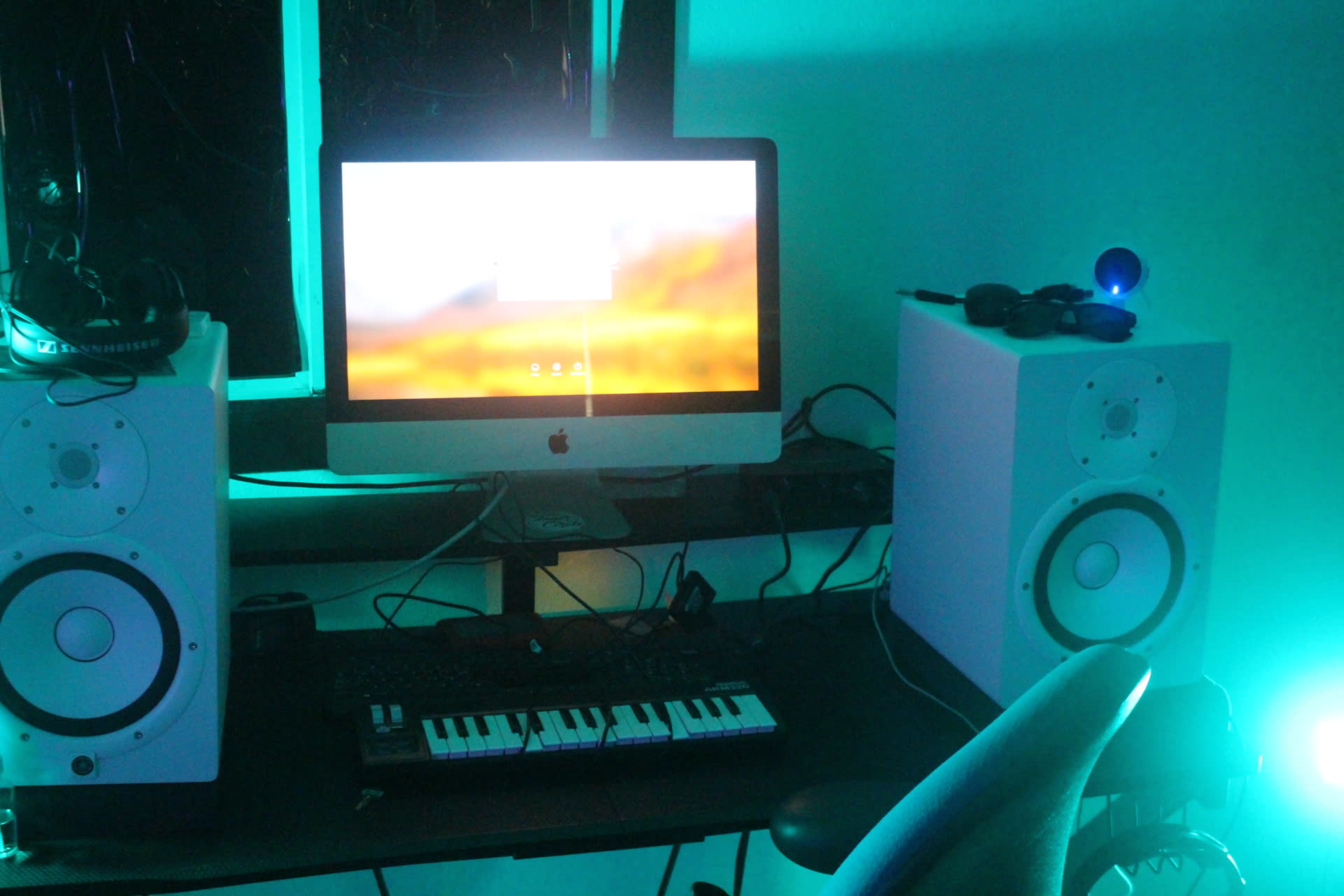 A desk holds a computer and two speakers, with a keyboard and headphones visible beside them, under blue lighting.