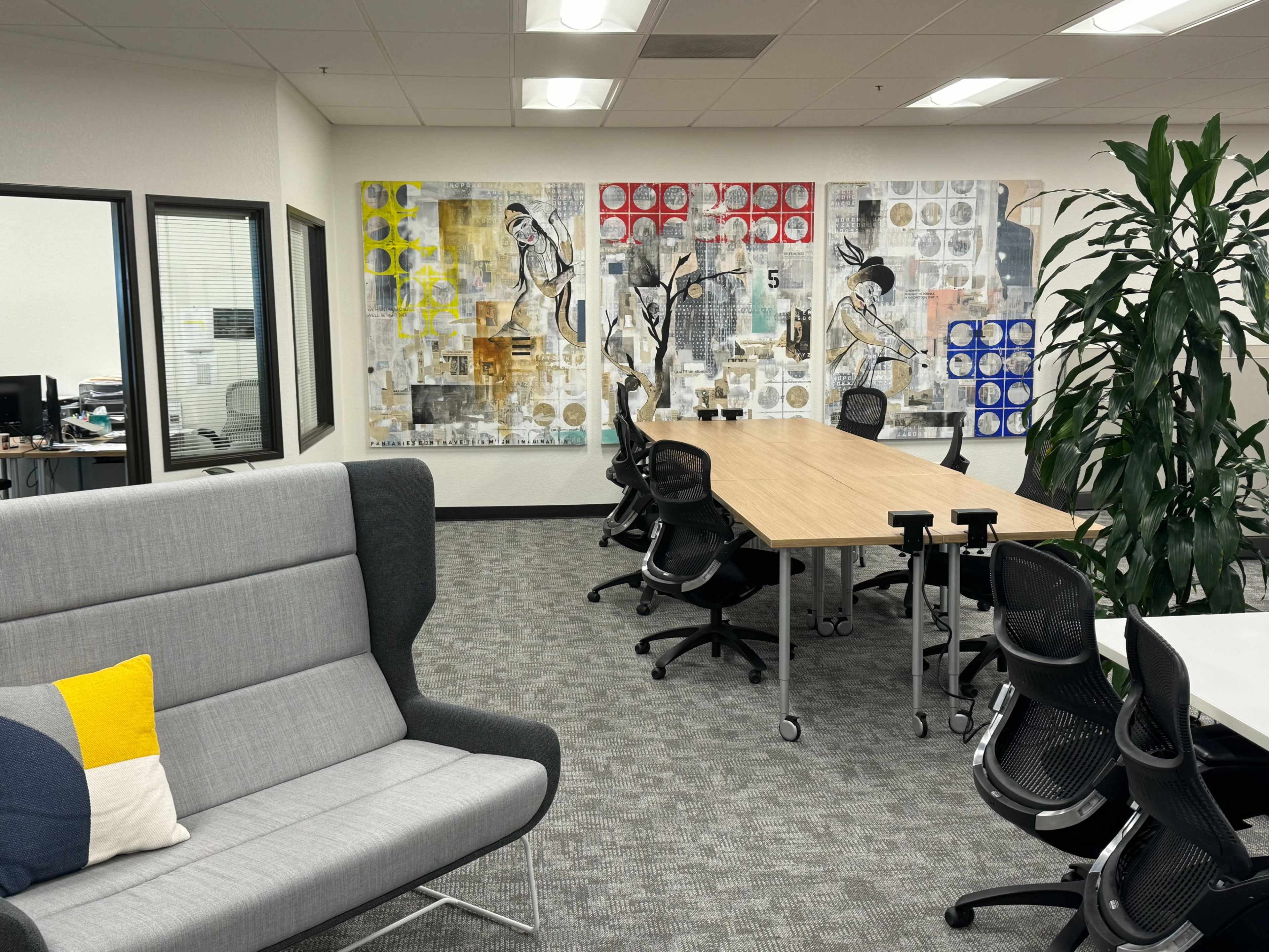 The image shows a modern office space featuring a large table surrounded by ergonomic chairs, a gray seating area, and abstract artwork on the walls.