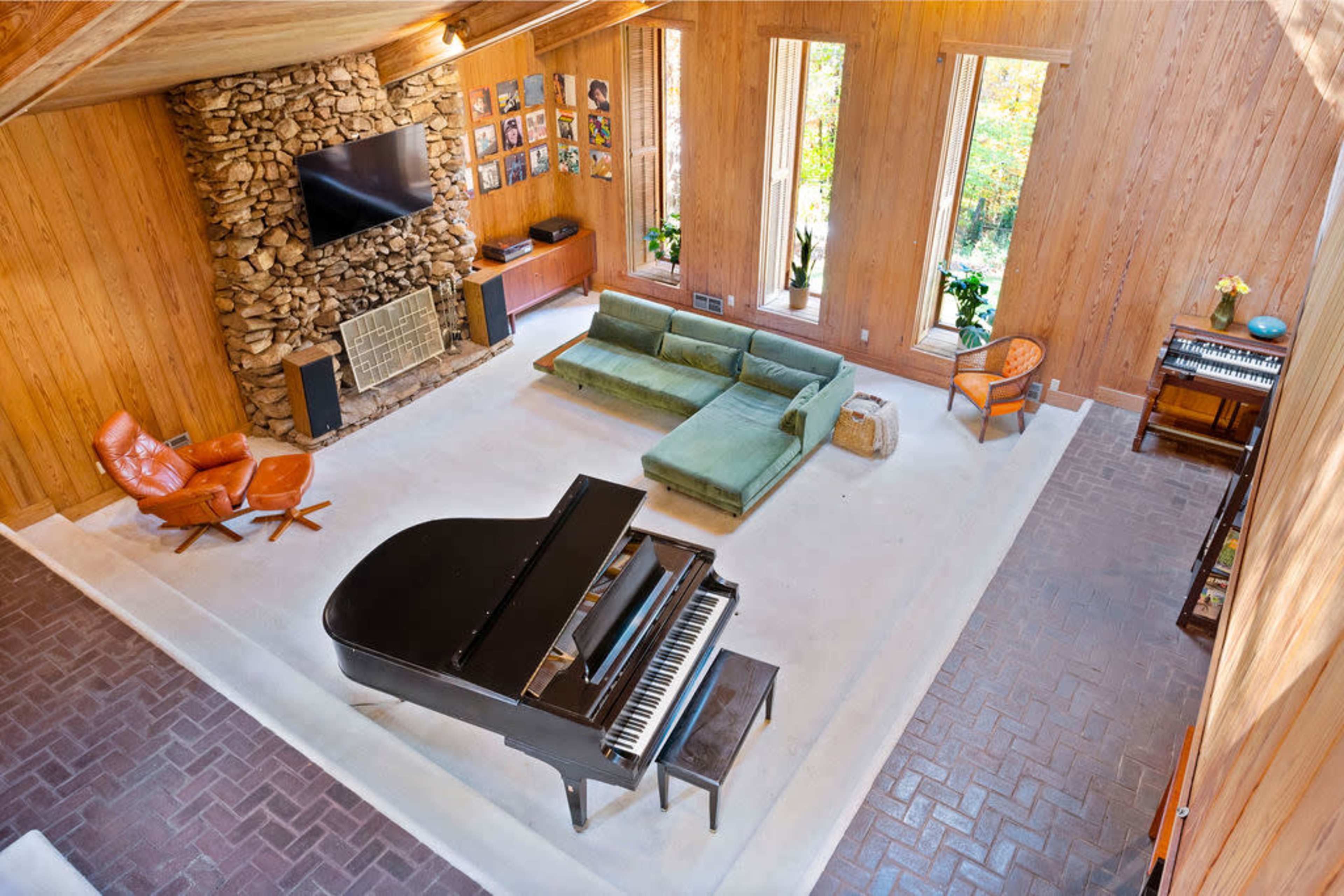 The room features a grand piano in the center, surrounded by a green sofa, two orange chairs, and wood-paneled walls, with large windows allowing natural light to enter.