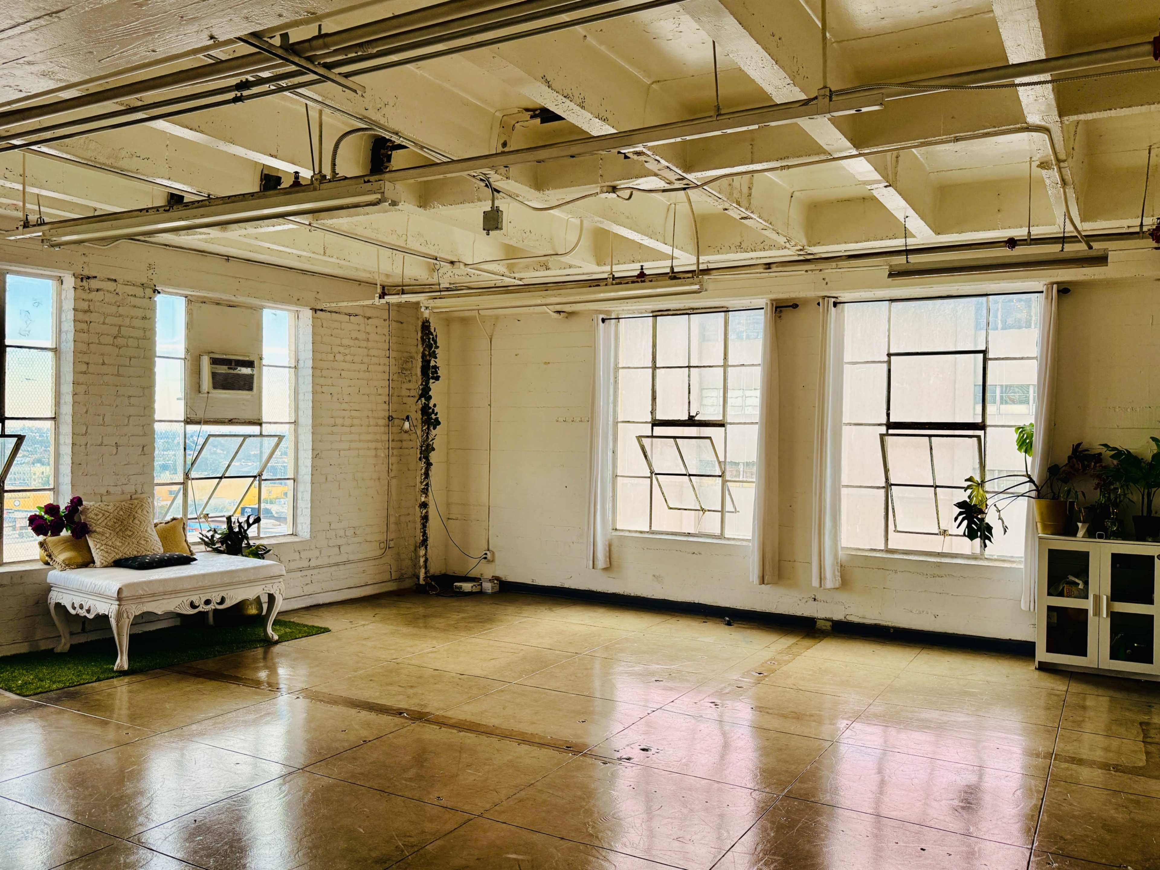 The image shows a spacious, well-lit room with large windows, a white sofa, and plants, featuring a minimalist design.