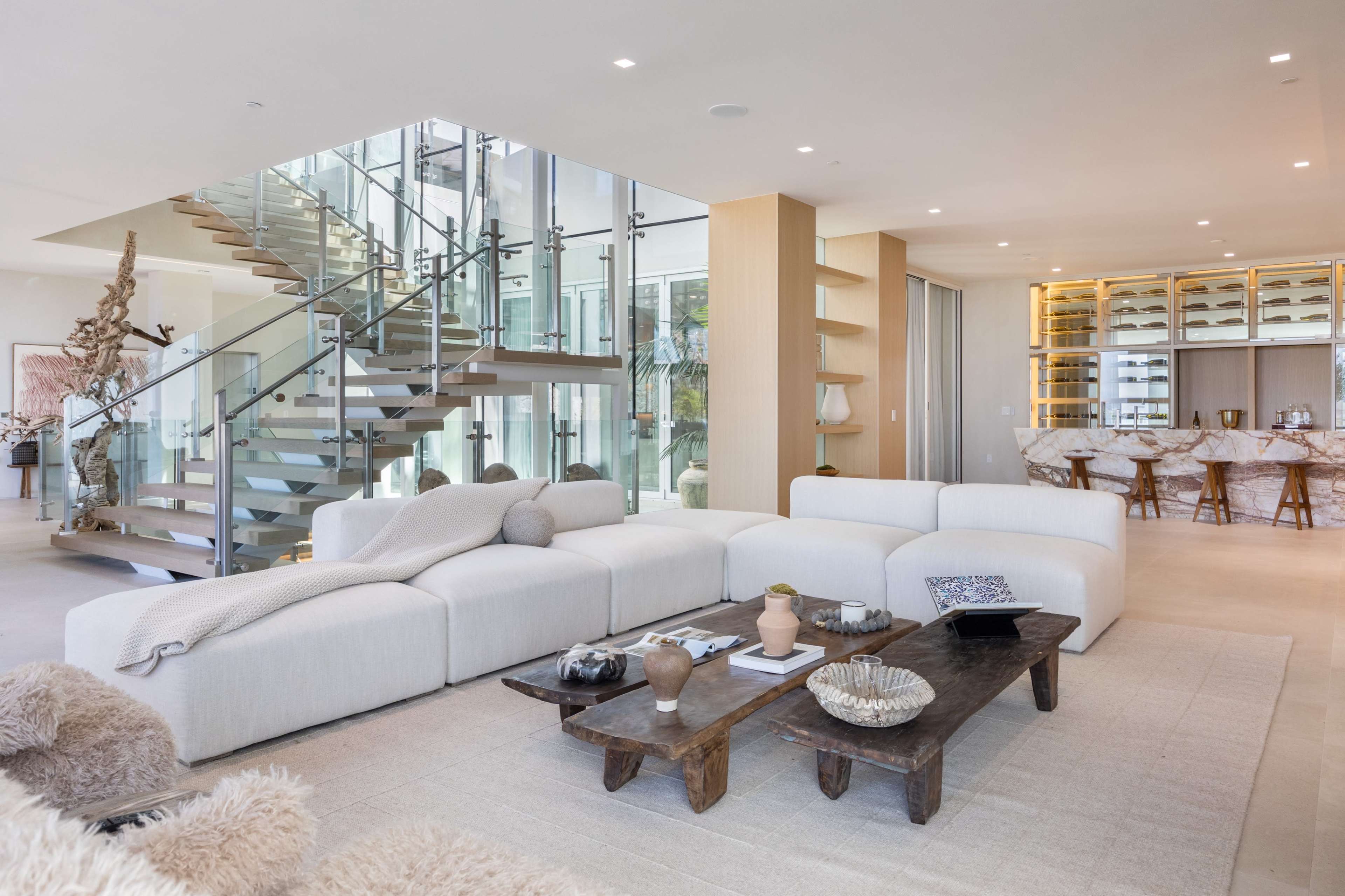 The image shows a modern, open-concept living area with a large sectional sofa, a wooden coffee table, and a staircase leading to an upper level, flanked by glass walls and a dining area in the background.