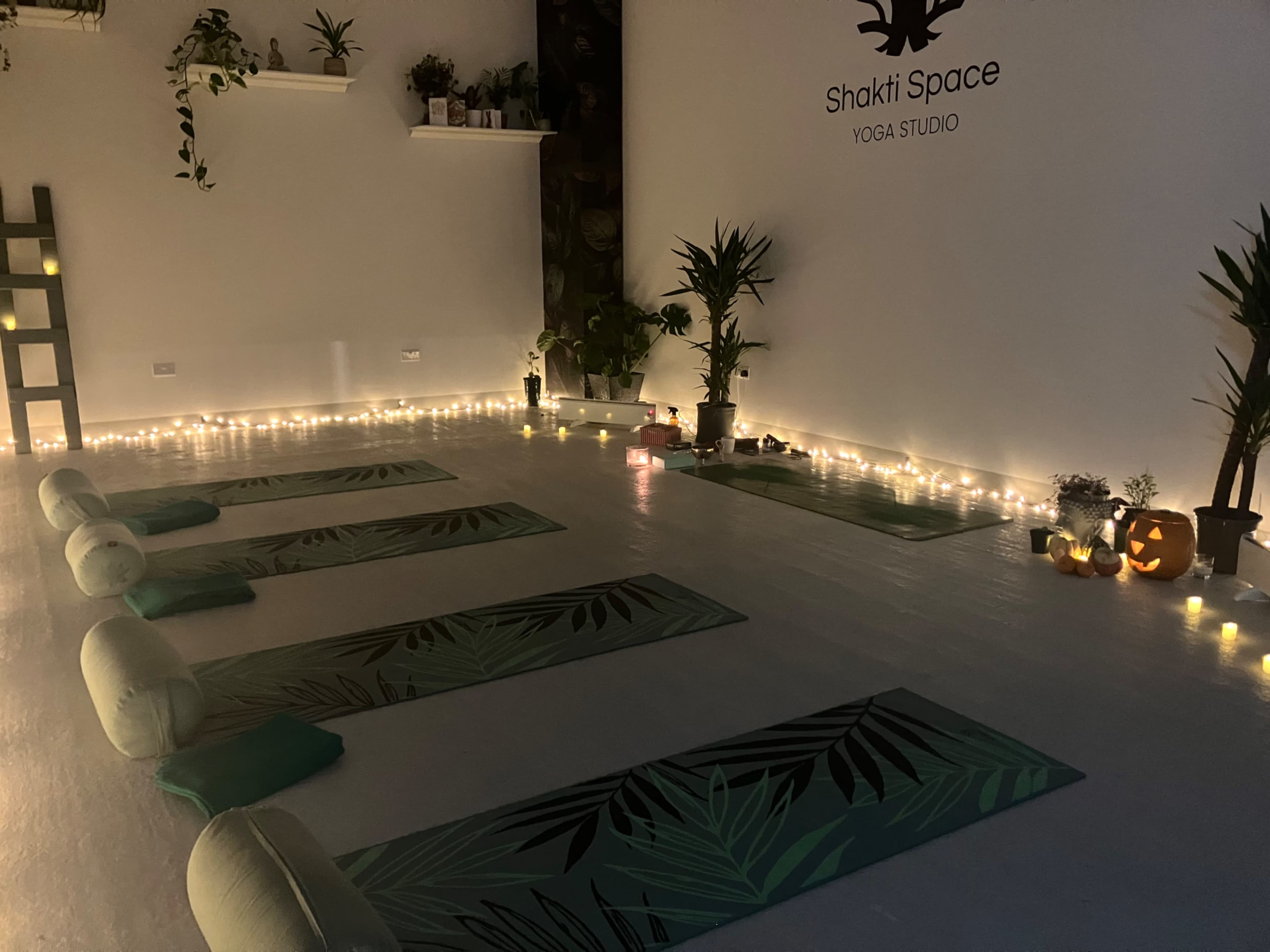 The image shows a yoga studio with yoga mats arranged neatly on the floor, surrounded by plants and soft lighting from candles.