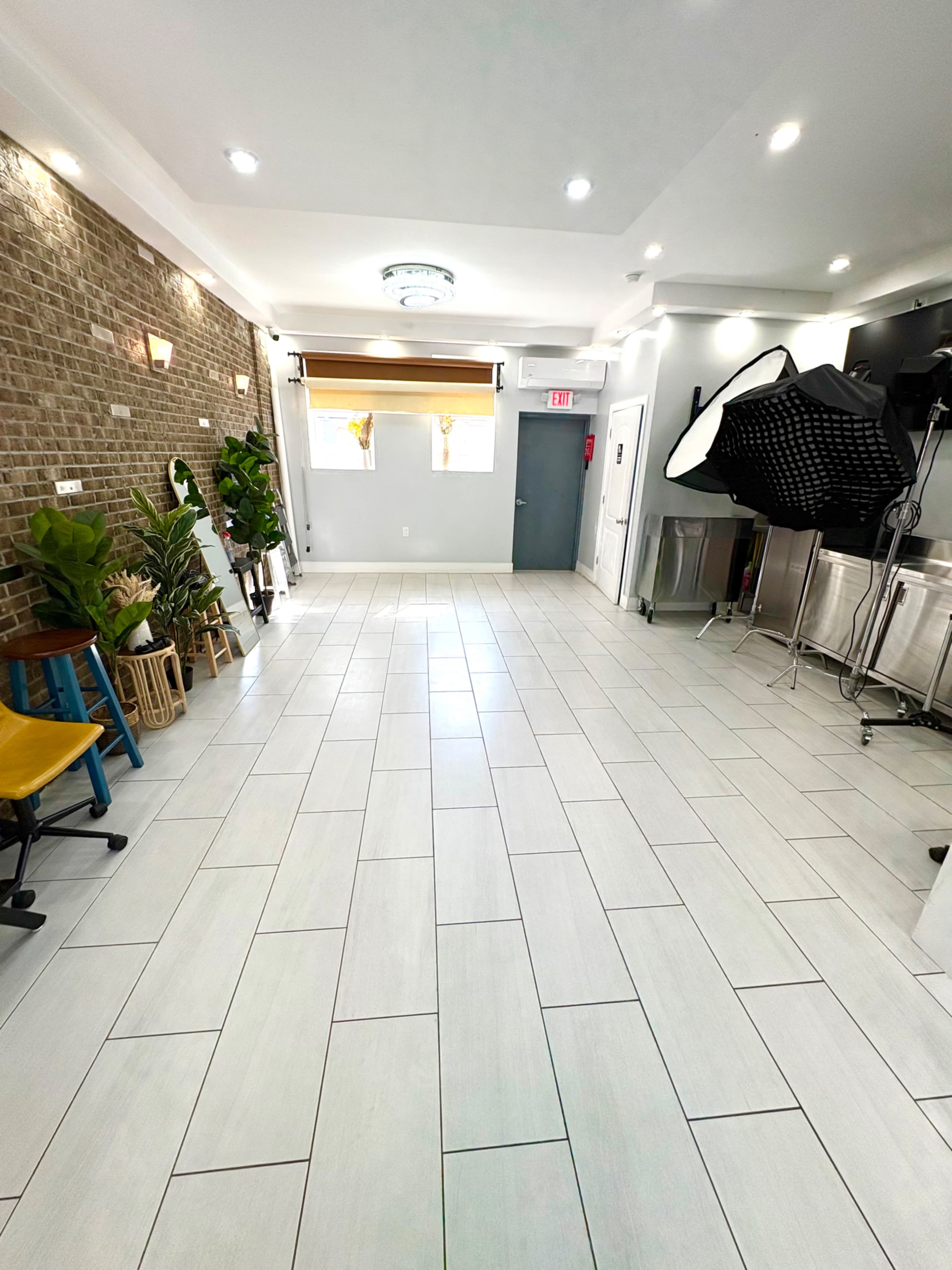 The image shows a spacious, well-lit room with tiled flooring, plants along the walls, and photography equipment set up in one corner.