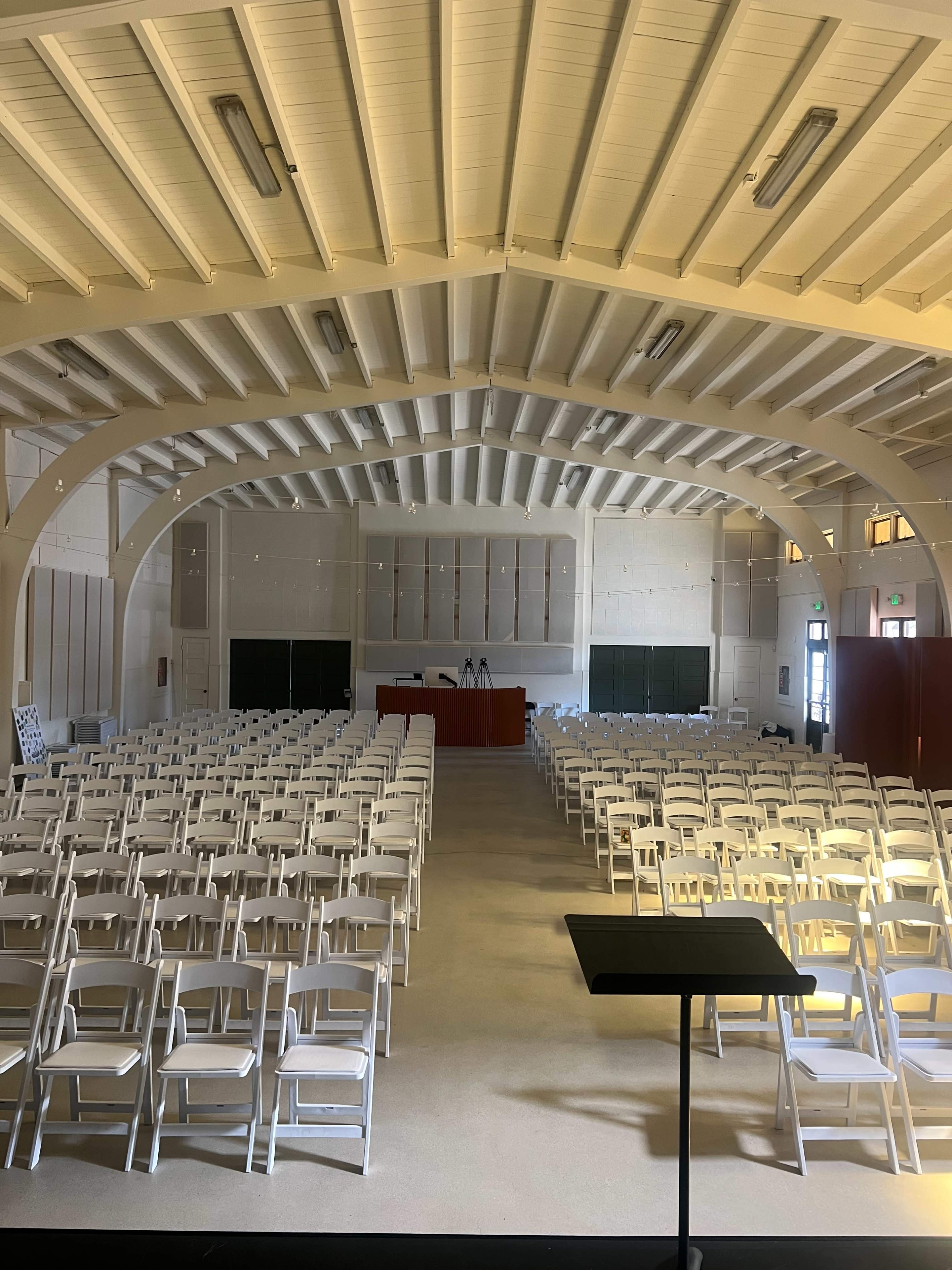 The image shows a large, empty auditorium with rows of white folding chairs and a stage at the front.