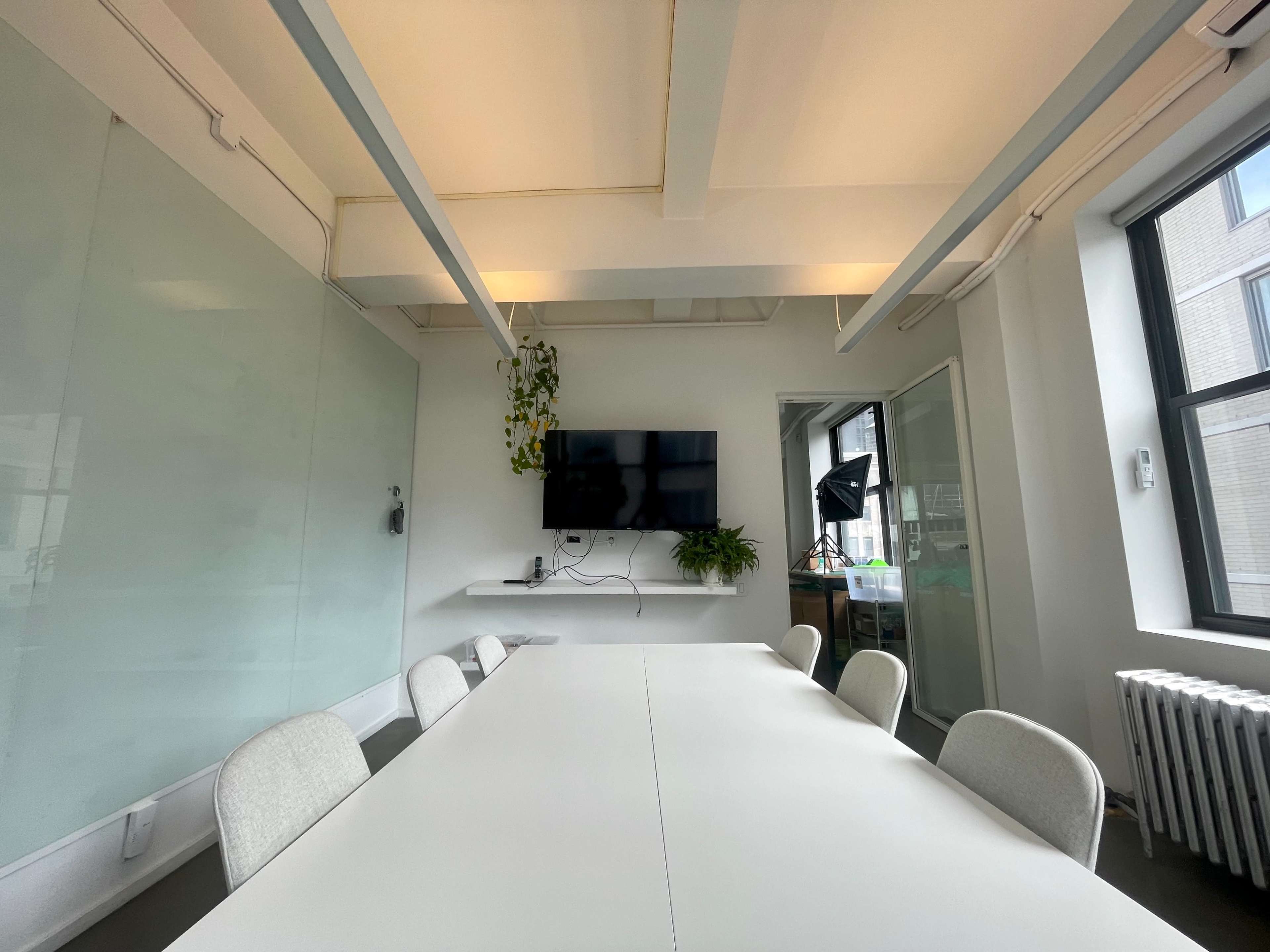 A bright conference room features a long white table, chairs, a television mounted on a wall, and large windows with a view of an urban environment.