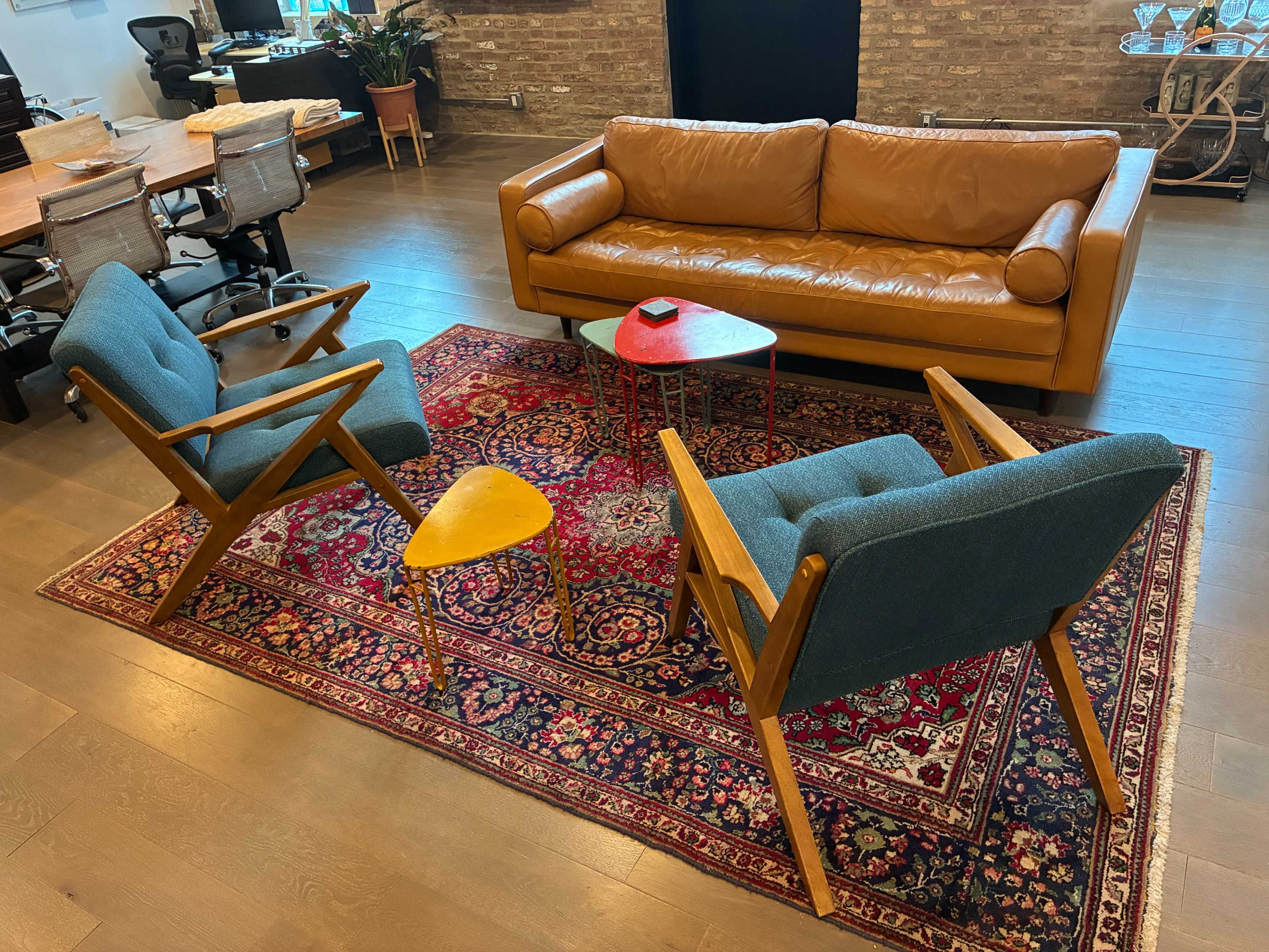 The image shows a living area with a brown leather sofa, two blue chairs, a small red side table, and a decorative rug on a wooden floor.