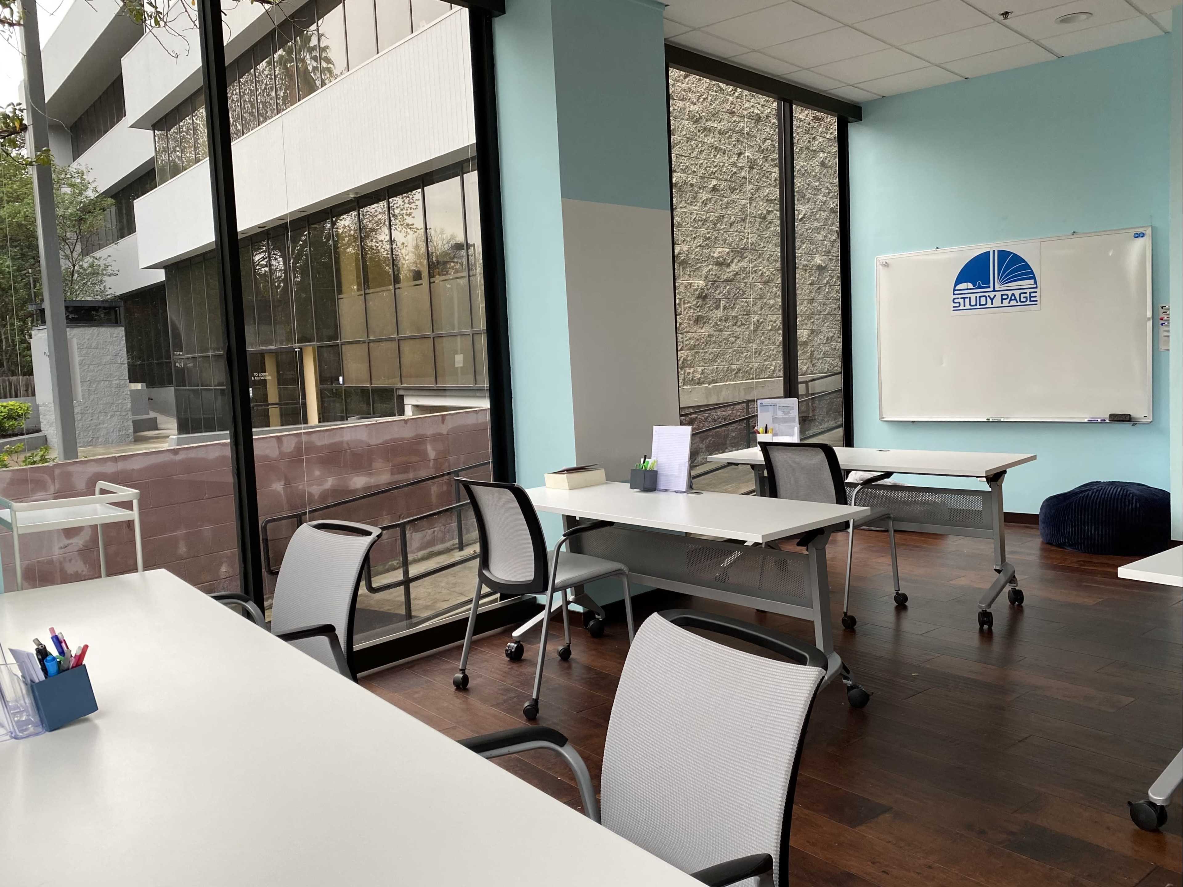 A bright study room with several white tables and chairs arranged near large windows, a whiteboard on the wall, and a few educational materials on the tables.