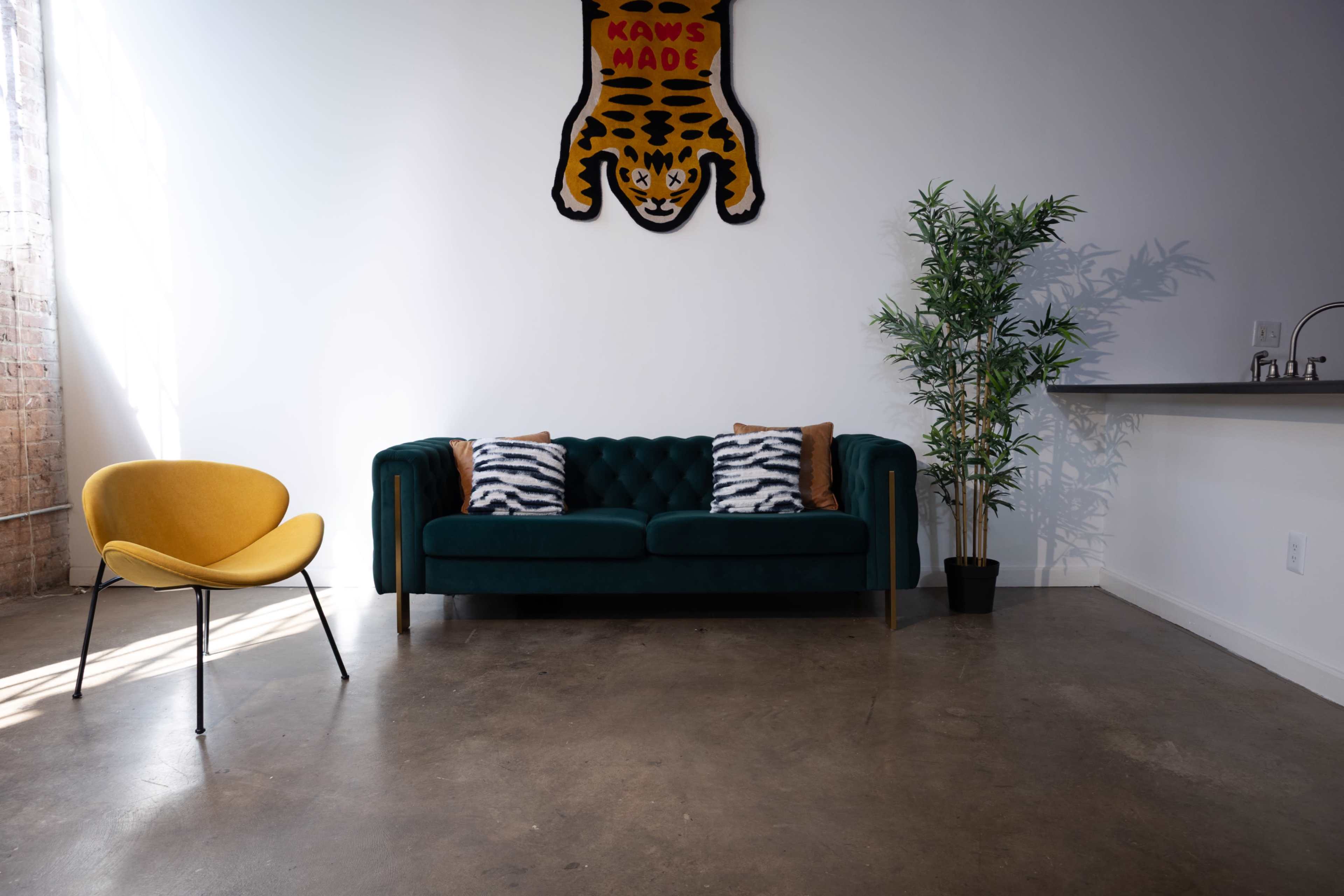 The image shows a modern interior featuring a green velvet sofa with decorative pillows, a yellow chair, a tall potted plant, and a wall hanging with a leopard design.