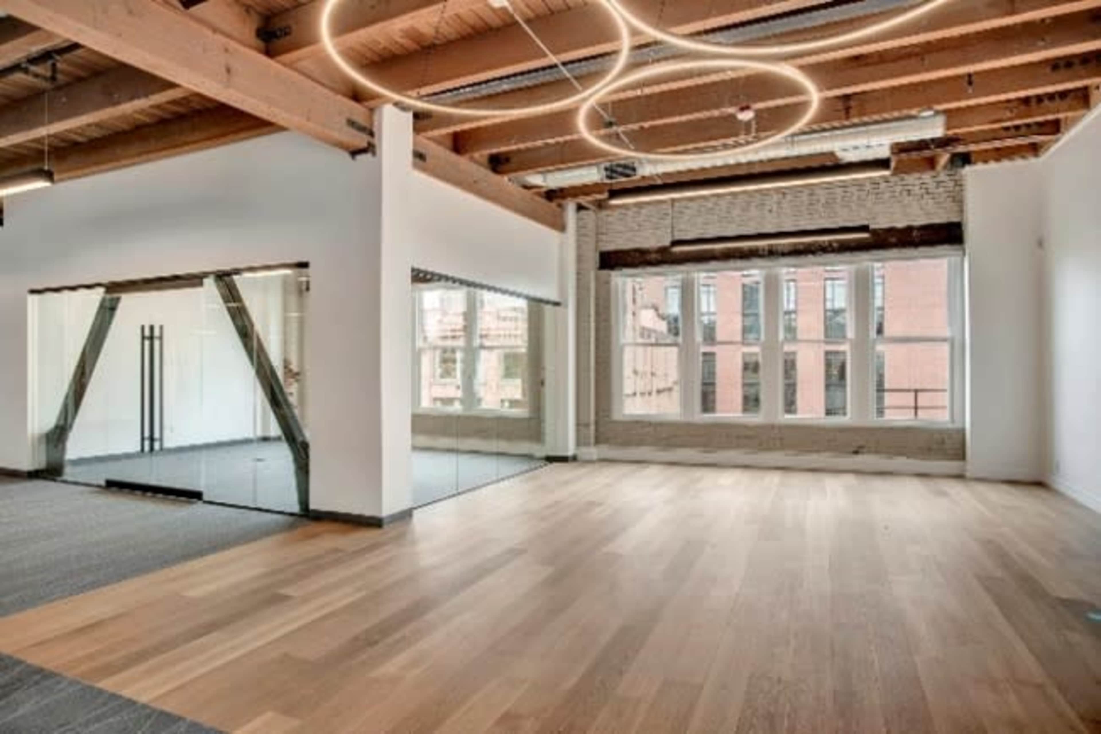 The image shows a spacious, modern room with wooden flooring, exposed beams, and large windows.