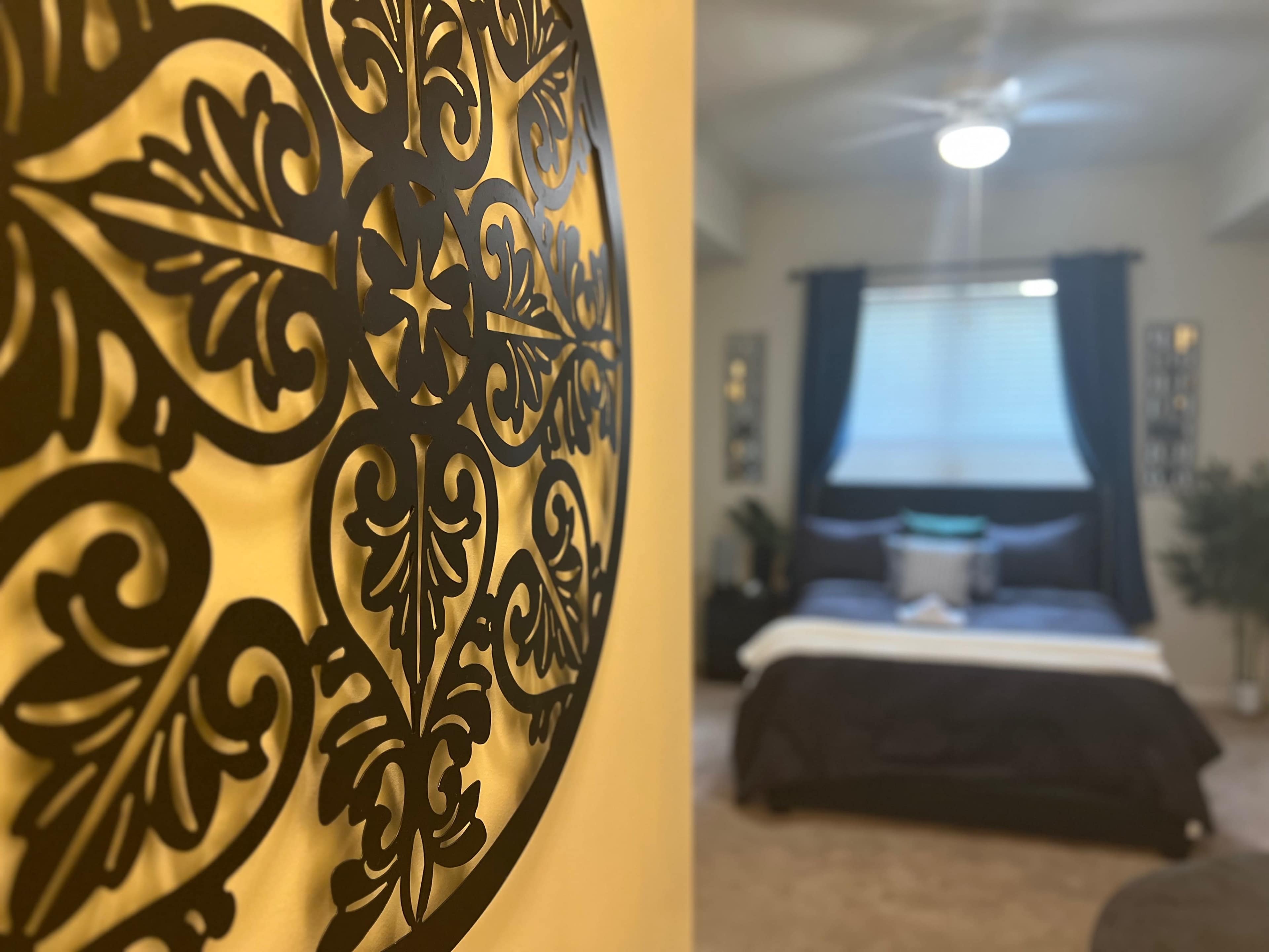 A decorative wall art piece is focused in the foreground, with a softly lit bedroom featuring a bed and window visible in the background.