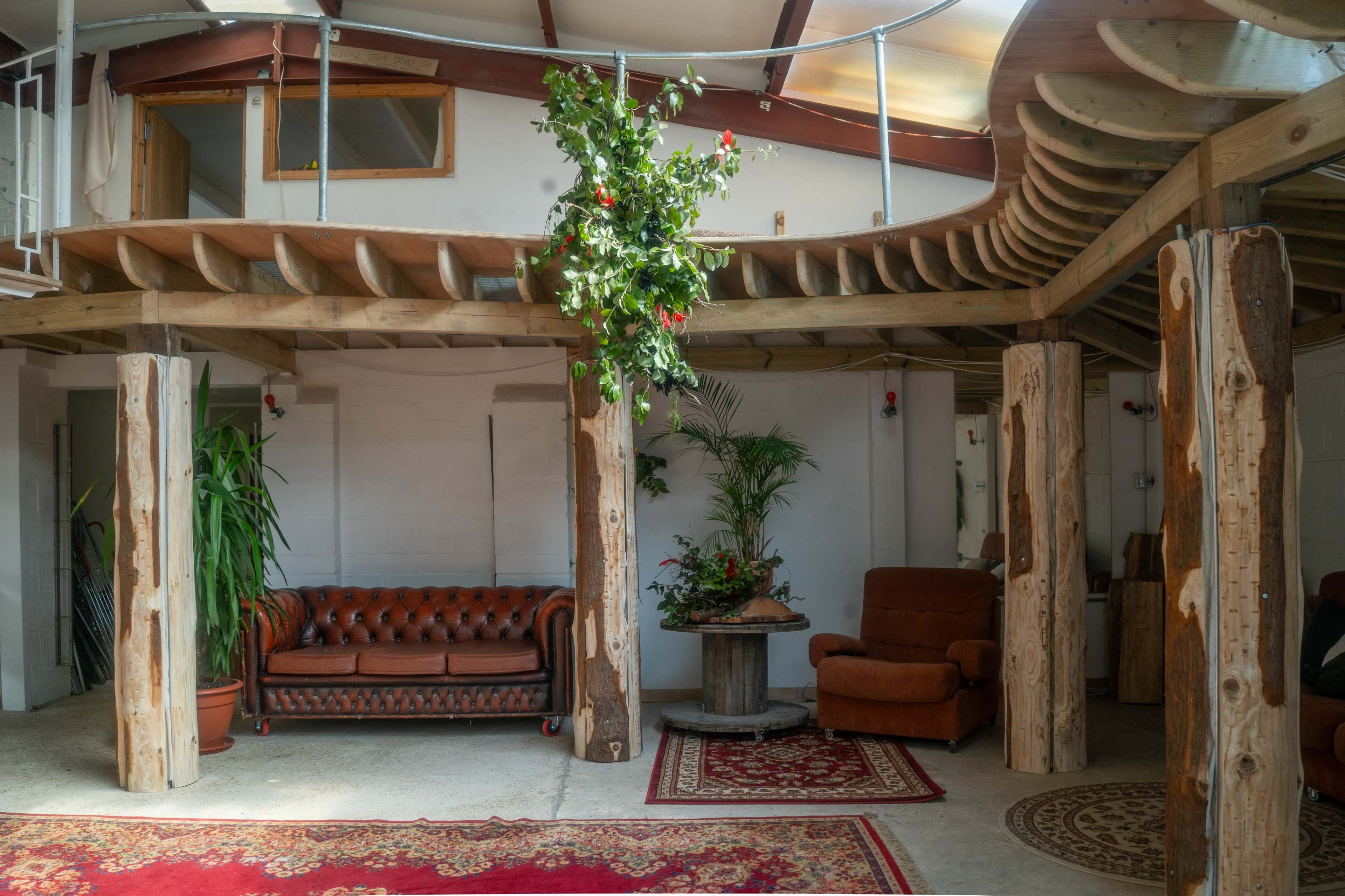 The image shows a spacious interior with wooden beams, a leather couch, two armchairs, and potted plants, complemented by a patterned rug.