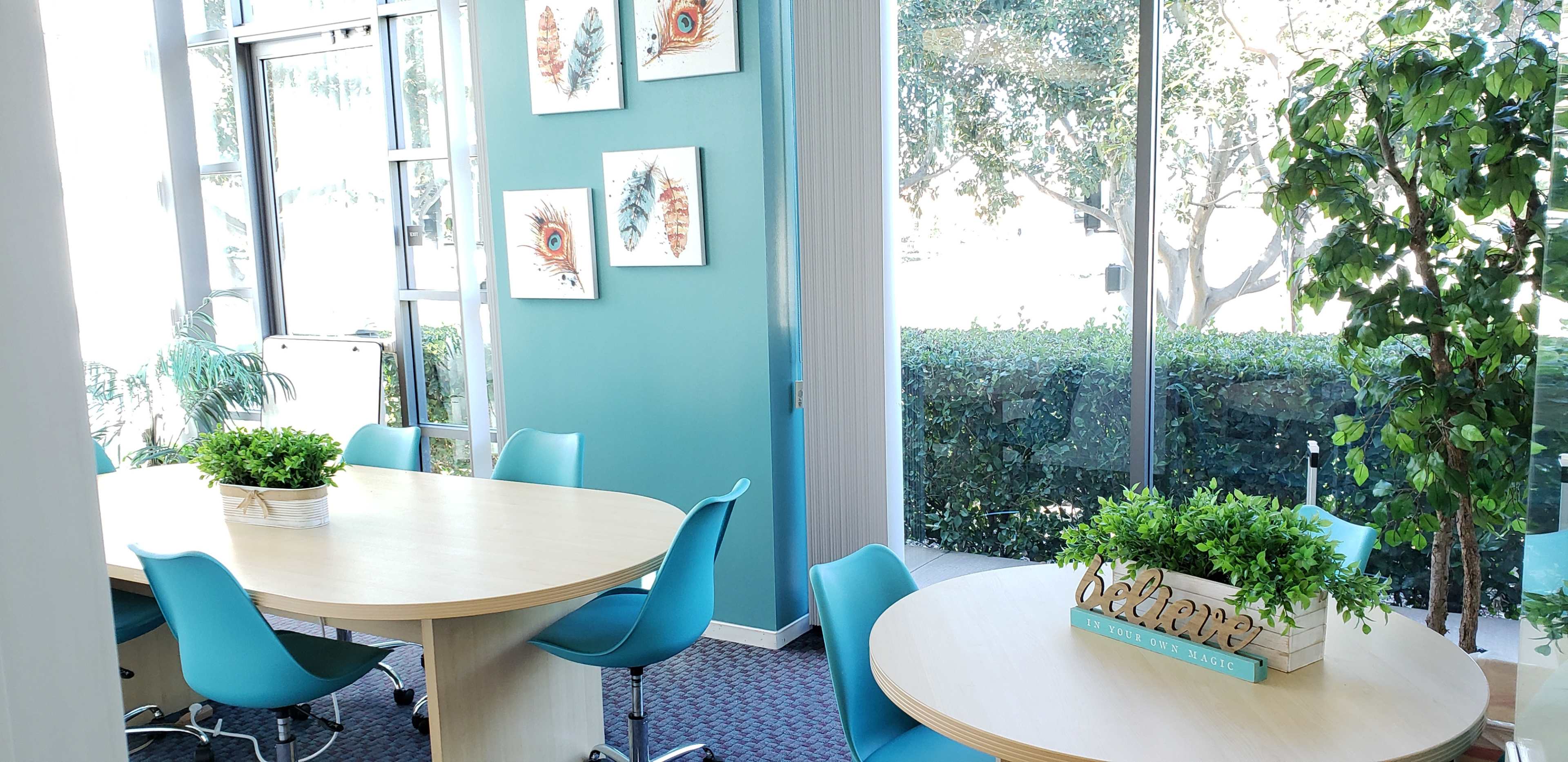 The image shows a bright meeting room with two round tables surrounded by blue chairs, decorated with potted plants and artwork on the walls.