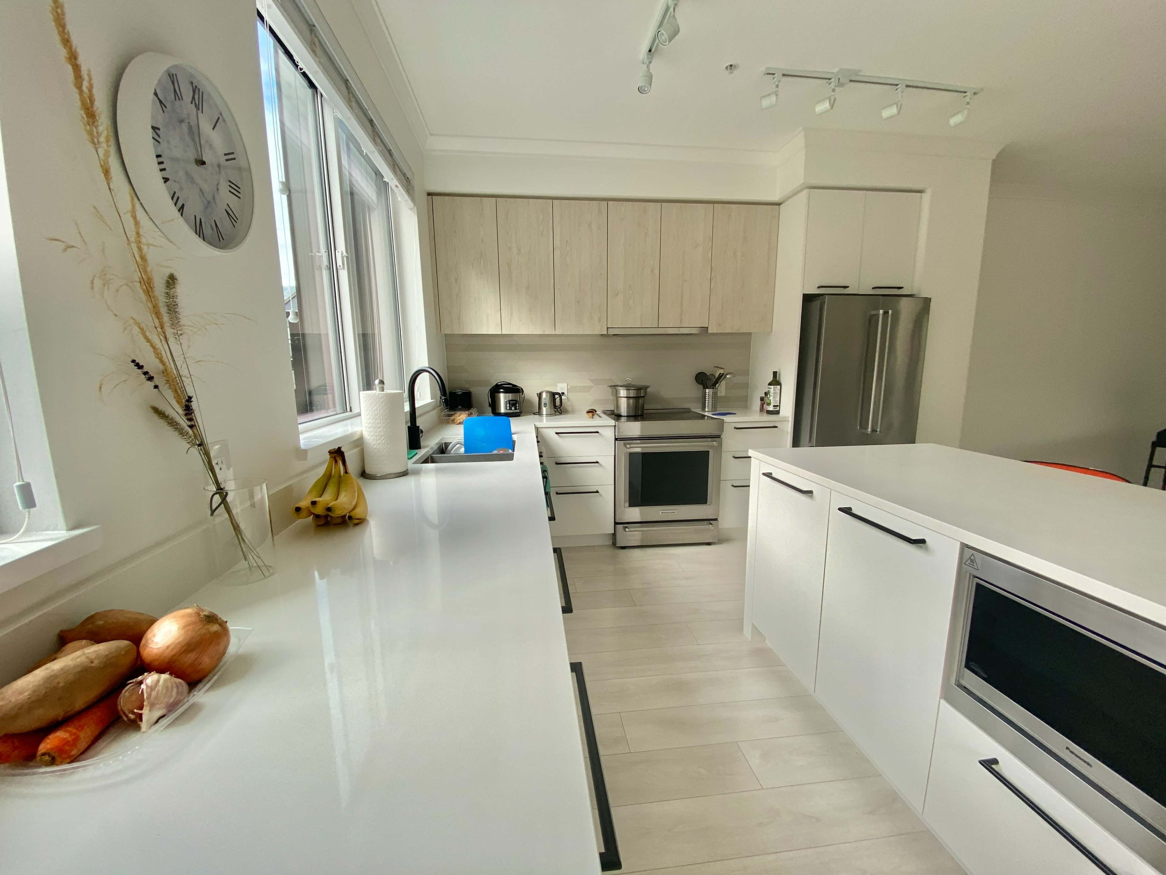 A modern kitchen features light wood cabinets, stainless steel appliances, and a white countertop with a bowl of fruits.