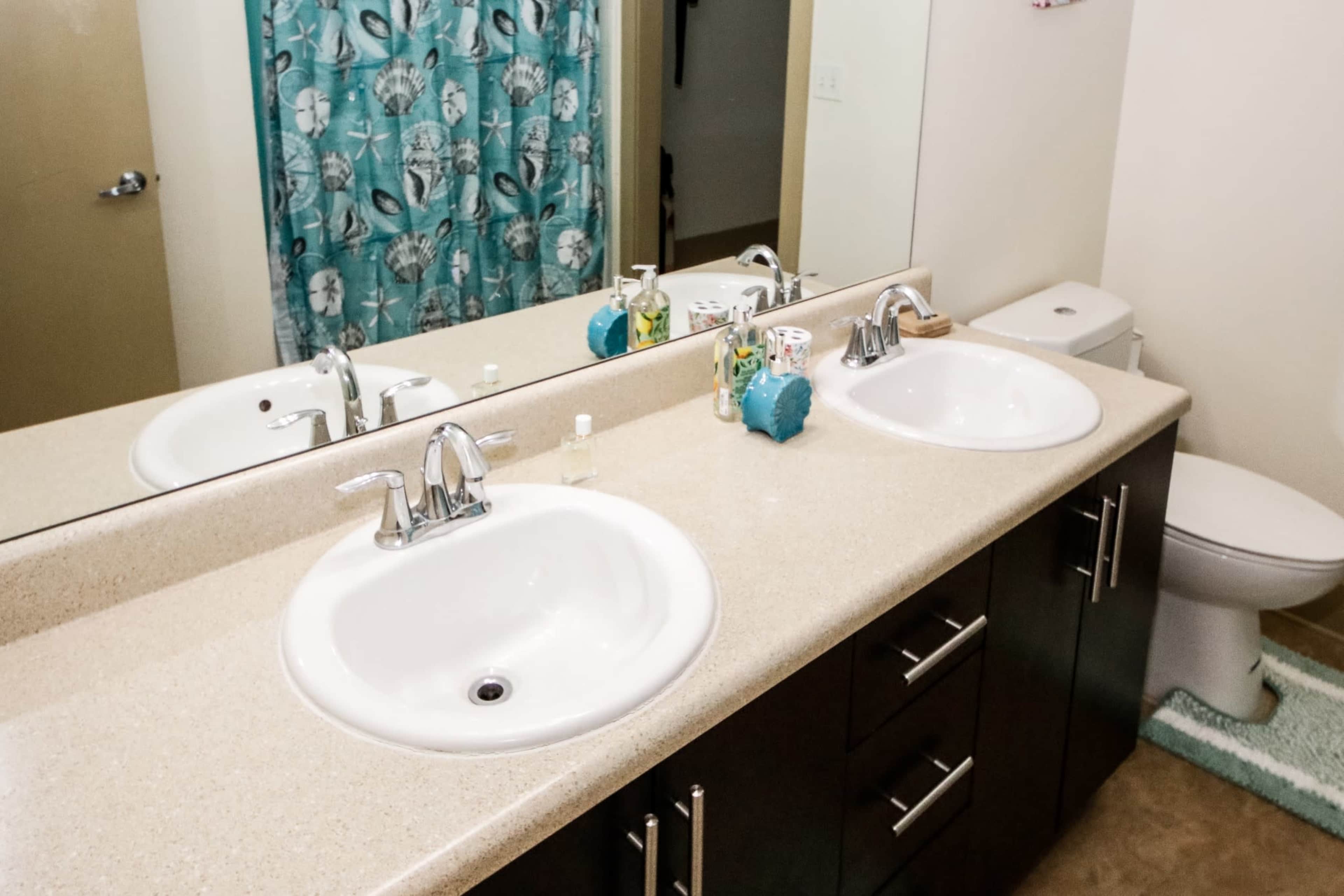 A bathroom features a dual sink vanity with a large mirror, a colorful shower curtain, and a toilet in the background.
