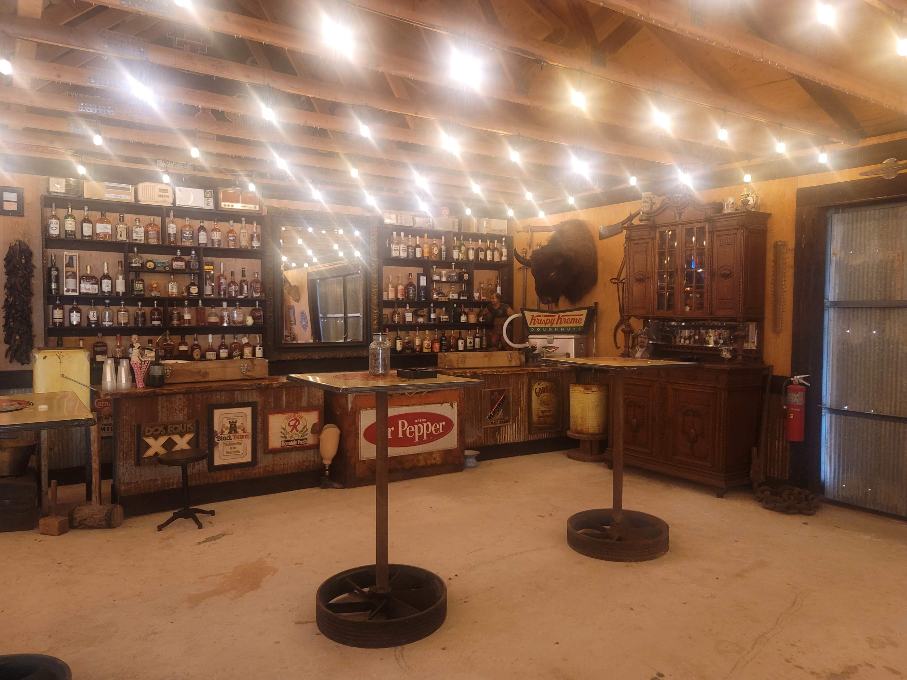 An interior of a rustic bar with shelves lined with various bottles, a wooden bar counter, and warm string lights illuminating the space.