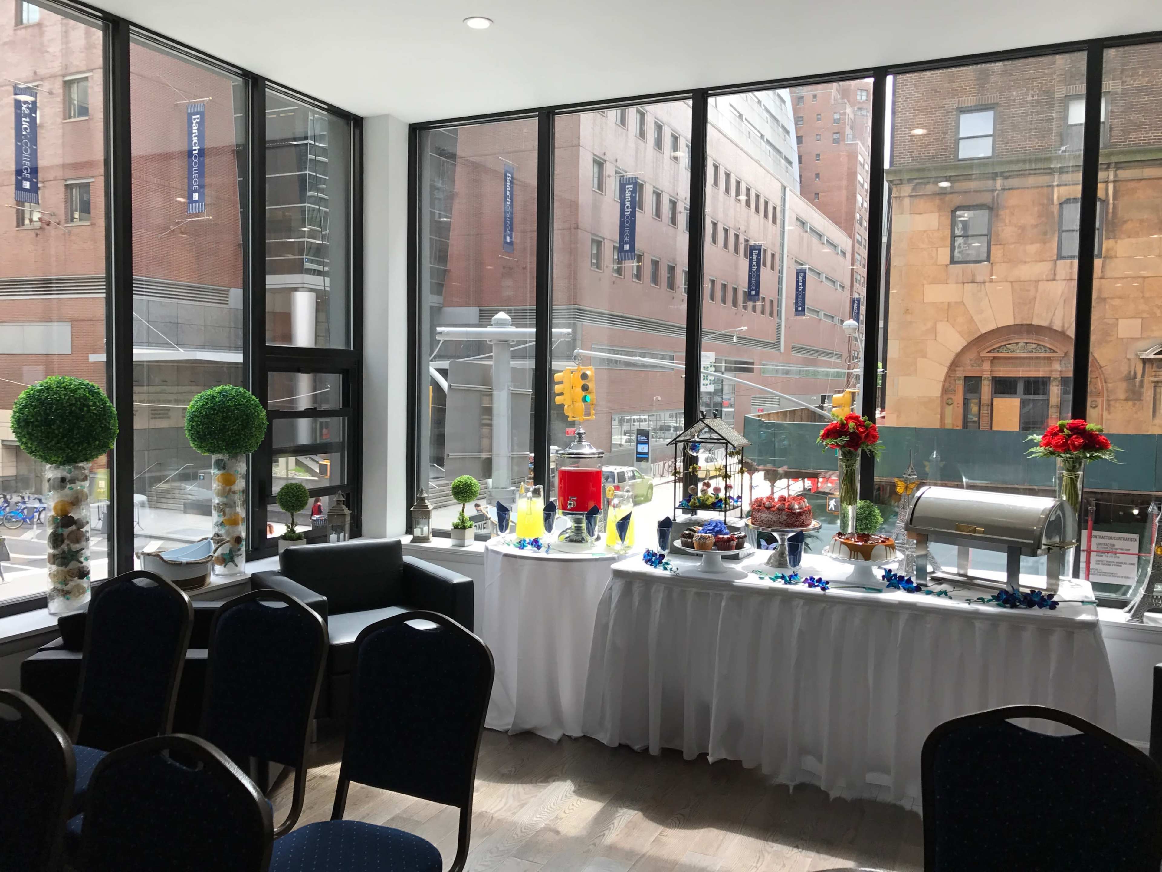 A buffet table is set up near large windows, displaying food and drink options, with greenery and flowers, overlooking a city street.