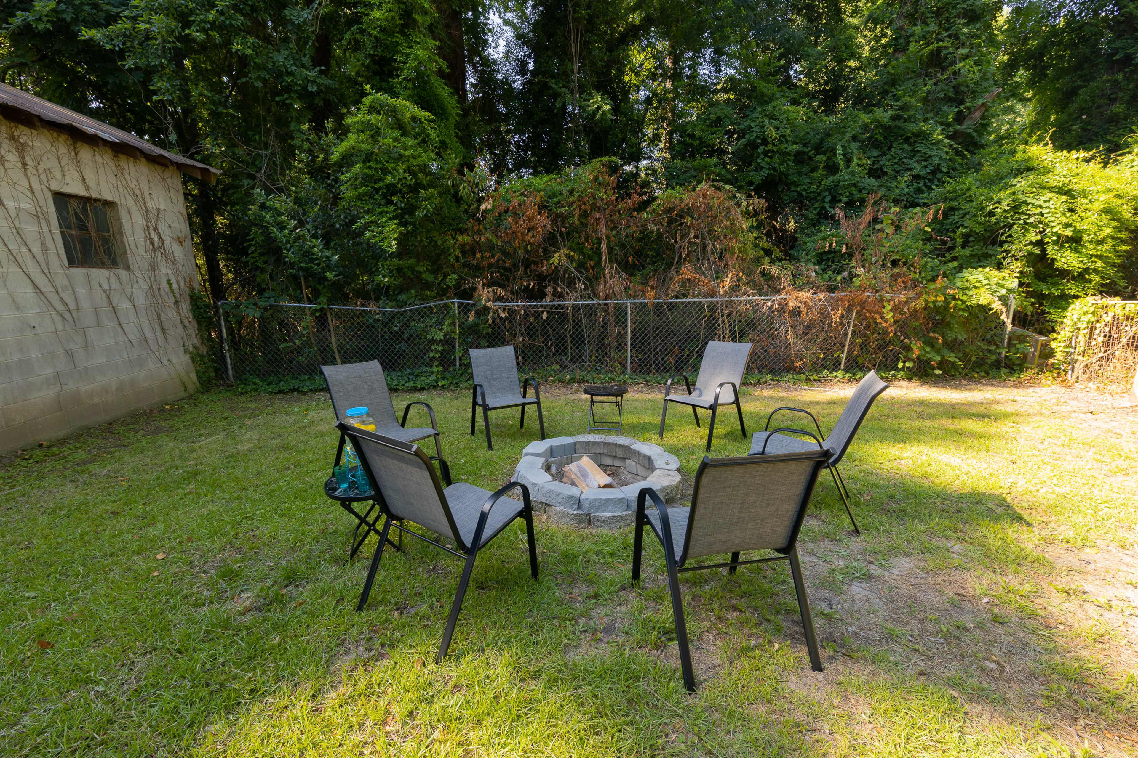 A circular stone fire pit is surrounded by six chairs in a grassy backyard, with a fence and trees in the background.