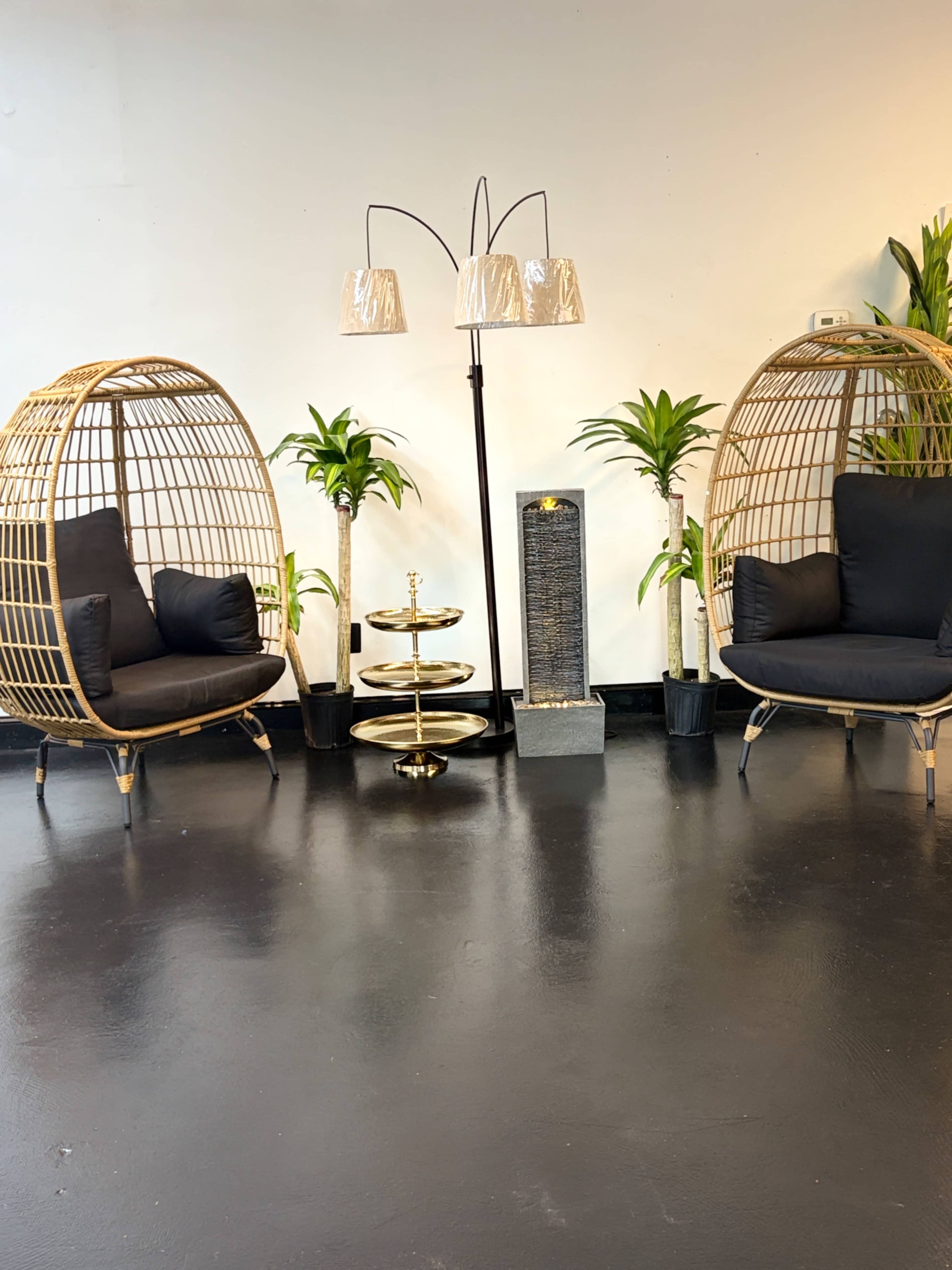 The image shows two rattan egg chairs positioned opposite each other, accompanied by a tall floor lamp and a tiered plant stand, all set against a plain wall.