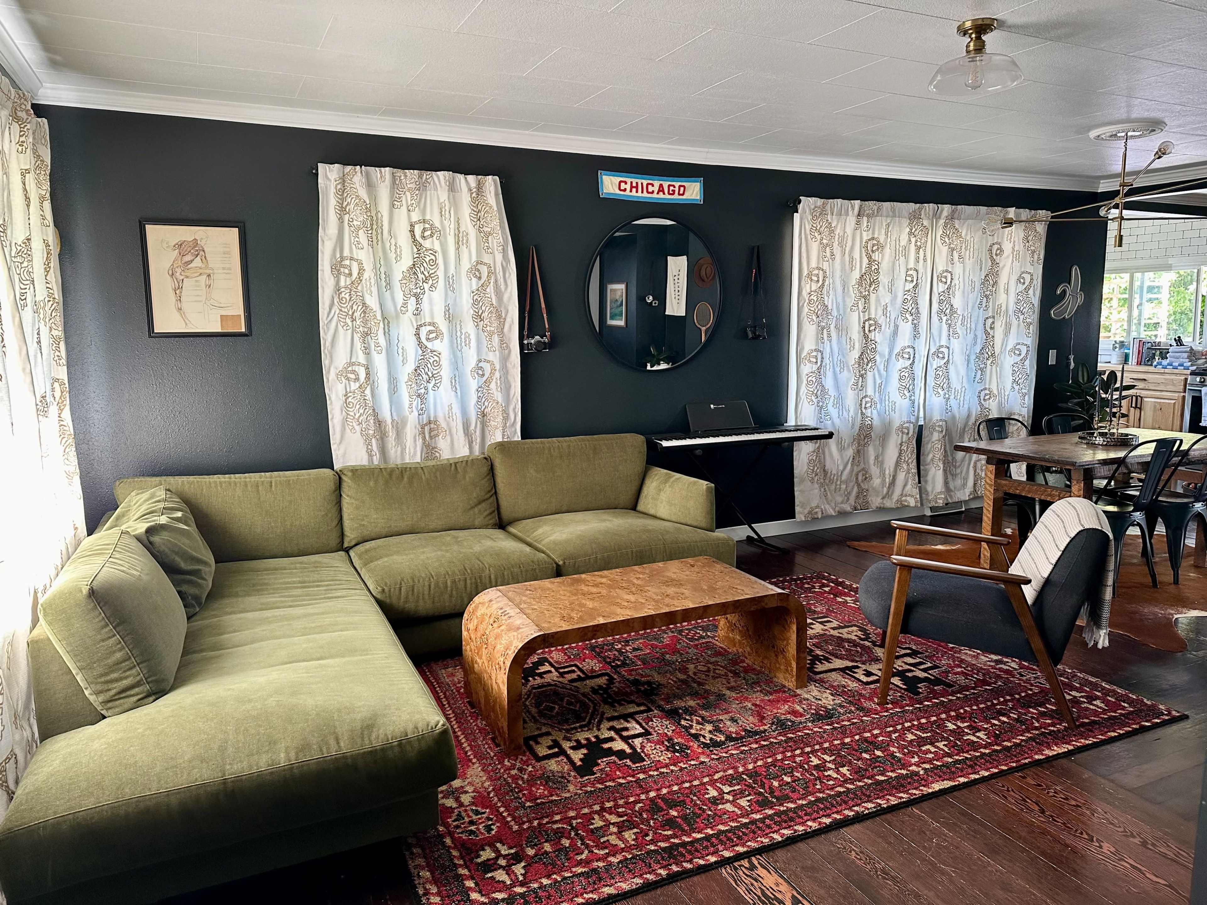 A living room with a green sectional sofa, a round mirror, and a wooden coffee table on a patterned rug, with literary art on the walls and curtains framing two windows.