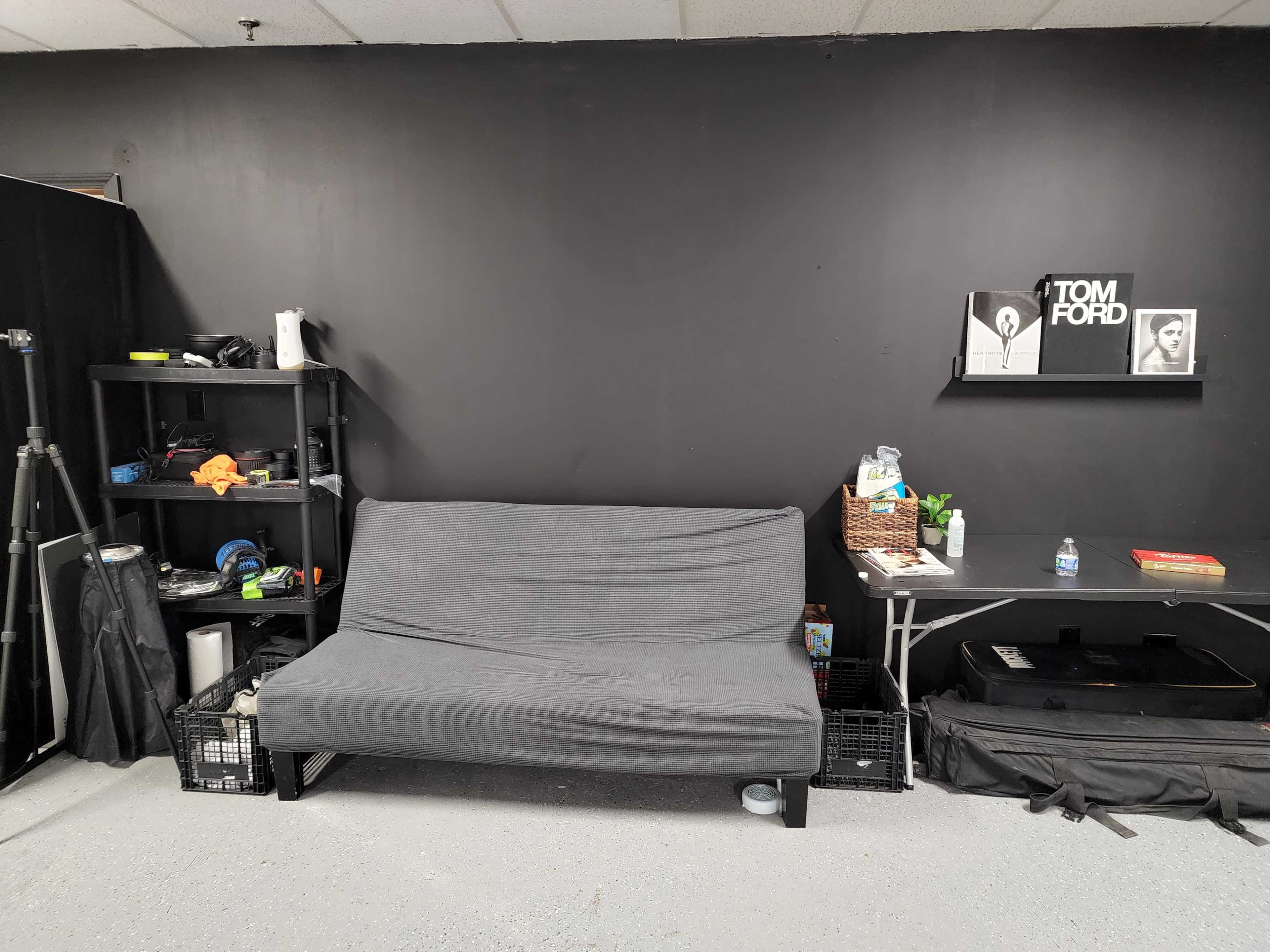 A minimalist interior featuring a gray futon against a black wall, with a shelf of items on one side and a table on the other holding a magazine and a basket.