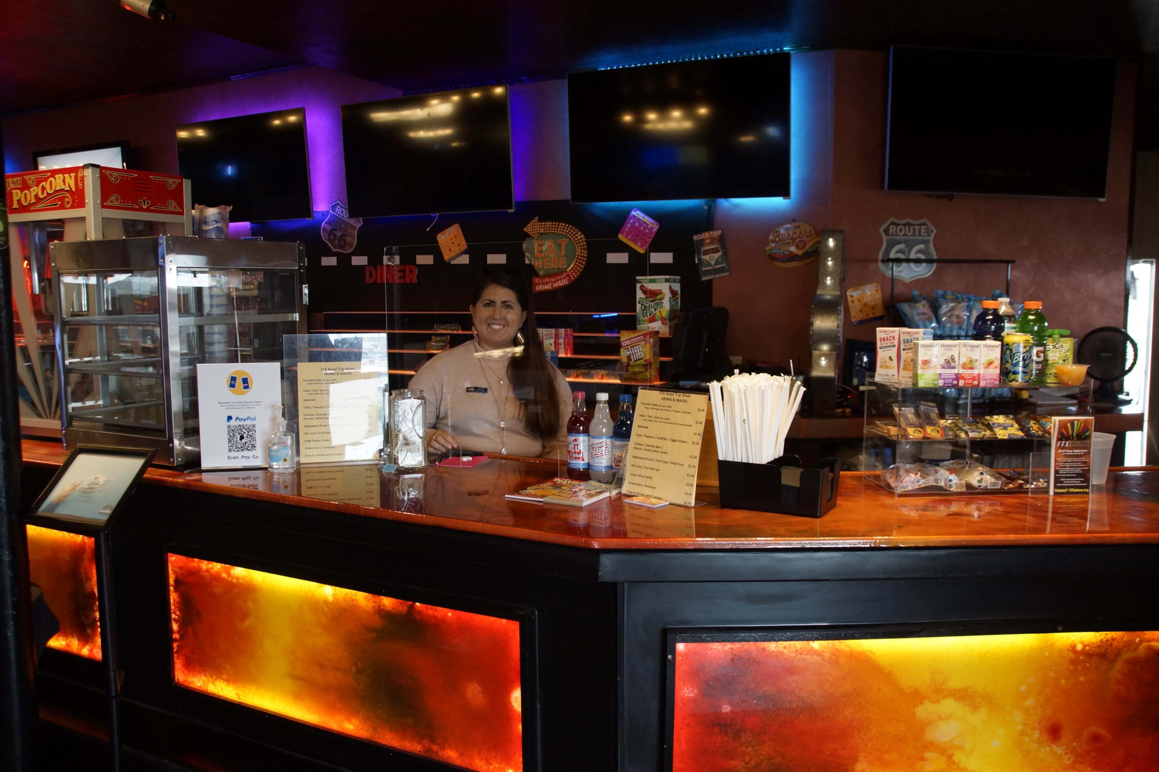 A woman stands behind the counter of a diner-style concession stand, surrounded by snacks and drinks.