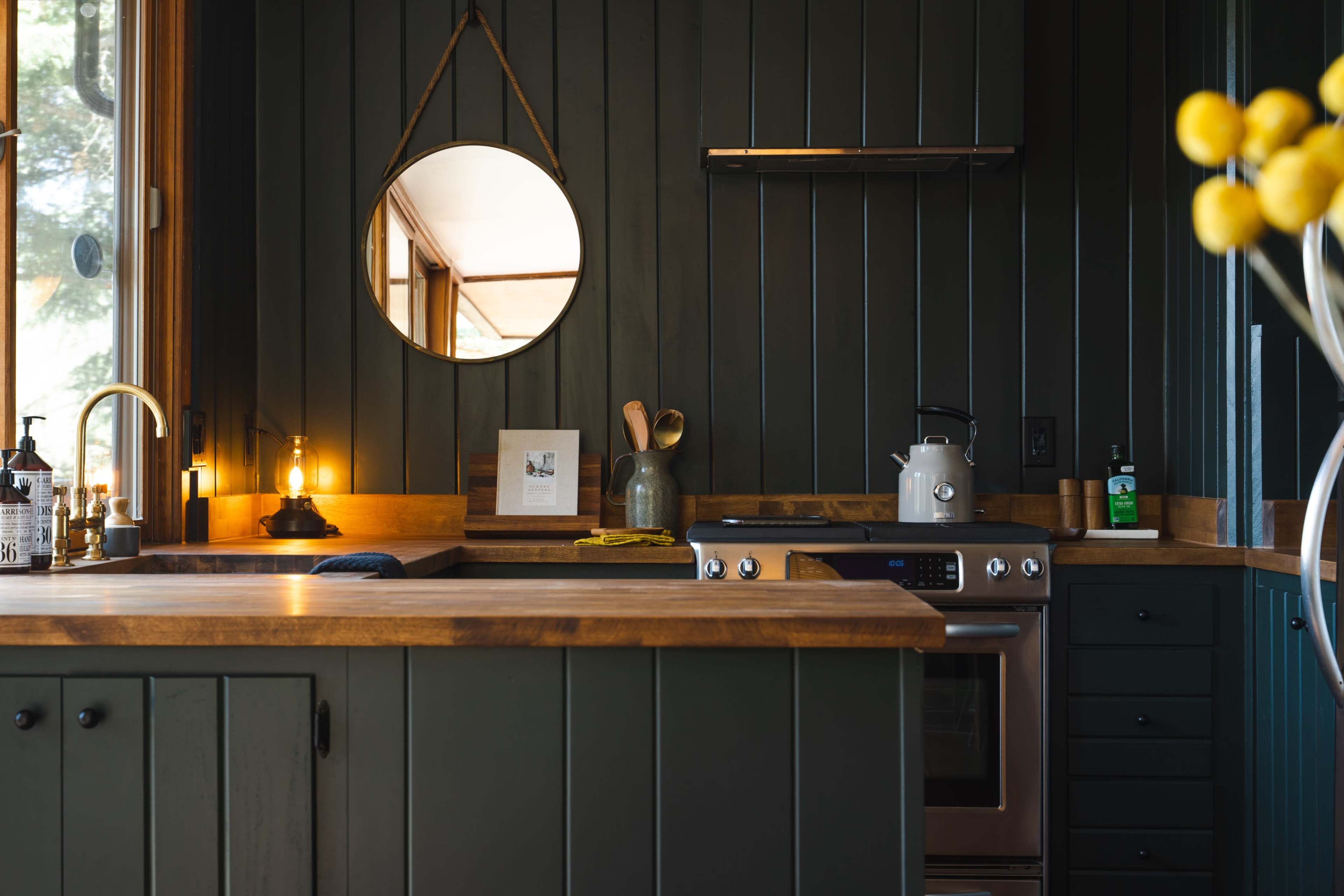 A modern kitchen with dark green paneling, a wooden countertop, and a round mirror mounted on the wall.