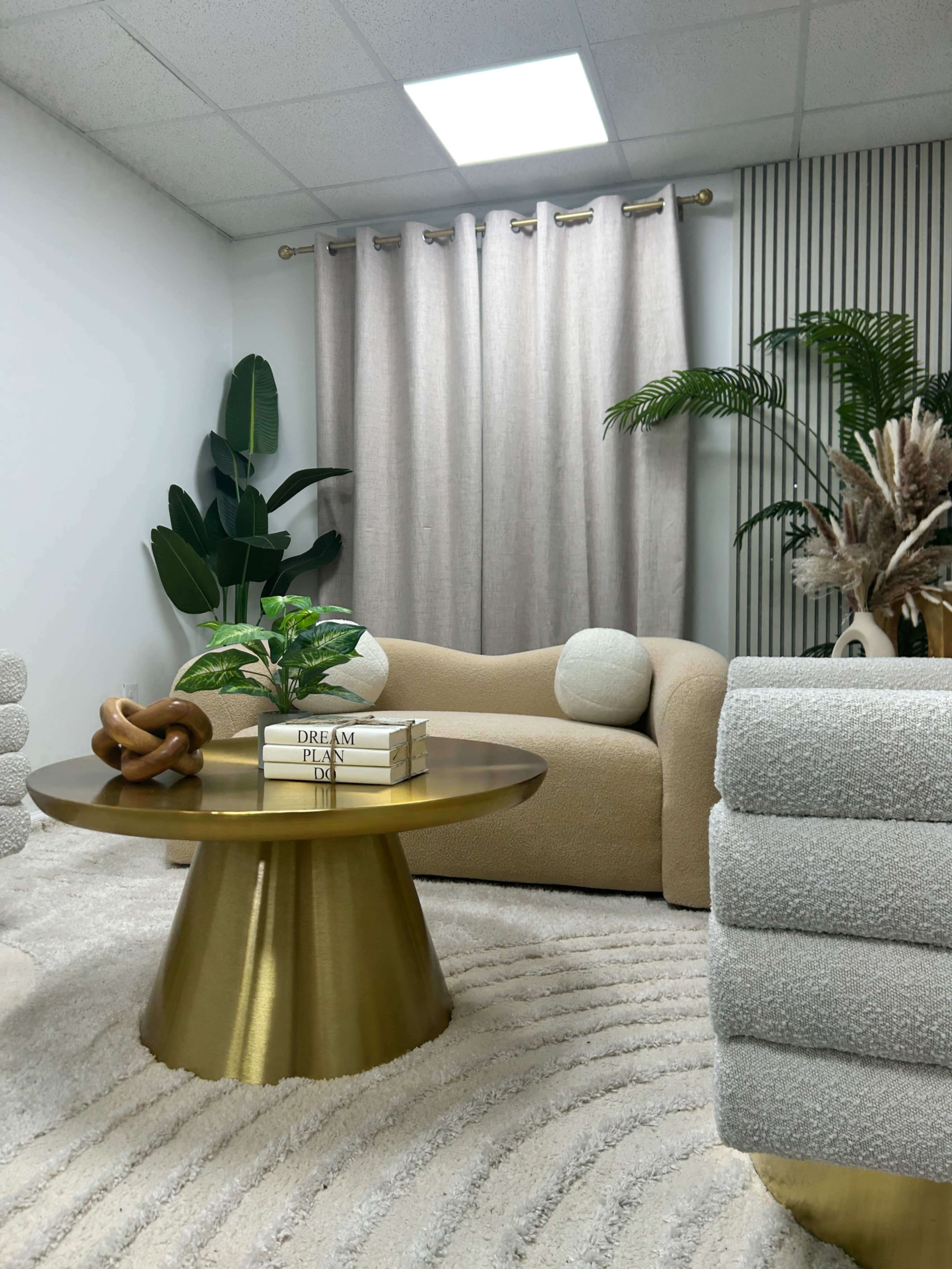 The image shows a modern interior with a light-colored sofa, a round gold coffee table, and decorative plants, set against a textured rug and draped curtains.
