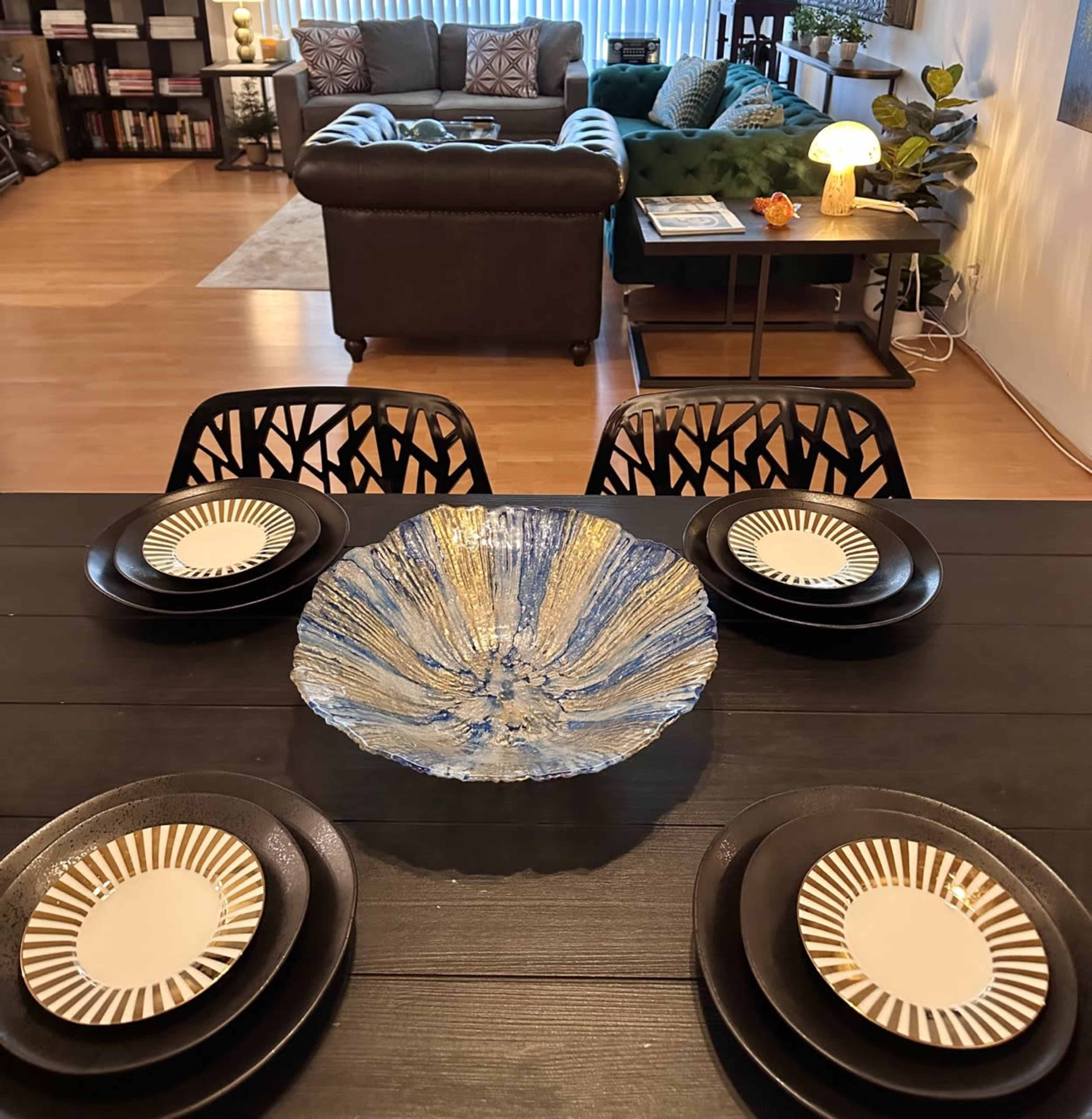 A dining table is set with black plates and a decorative blue bowl, overlooking a living room with a green couch and bookshelves.