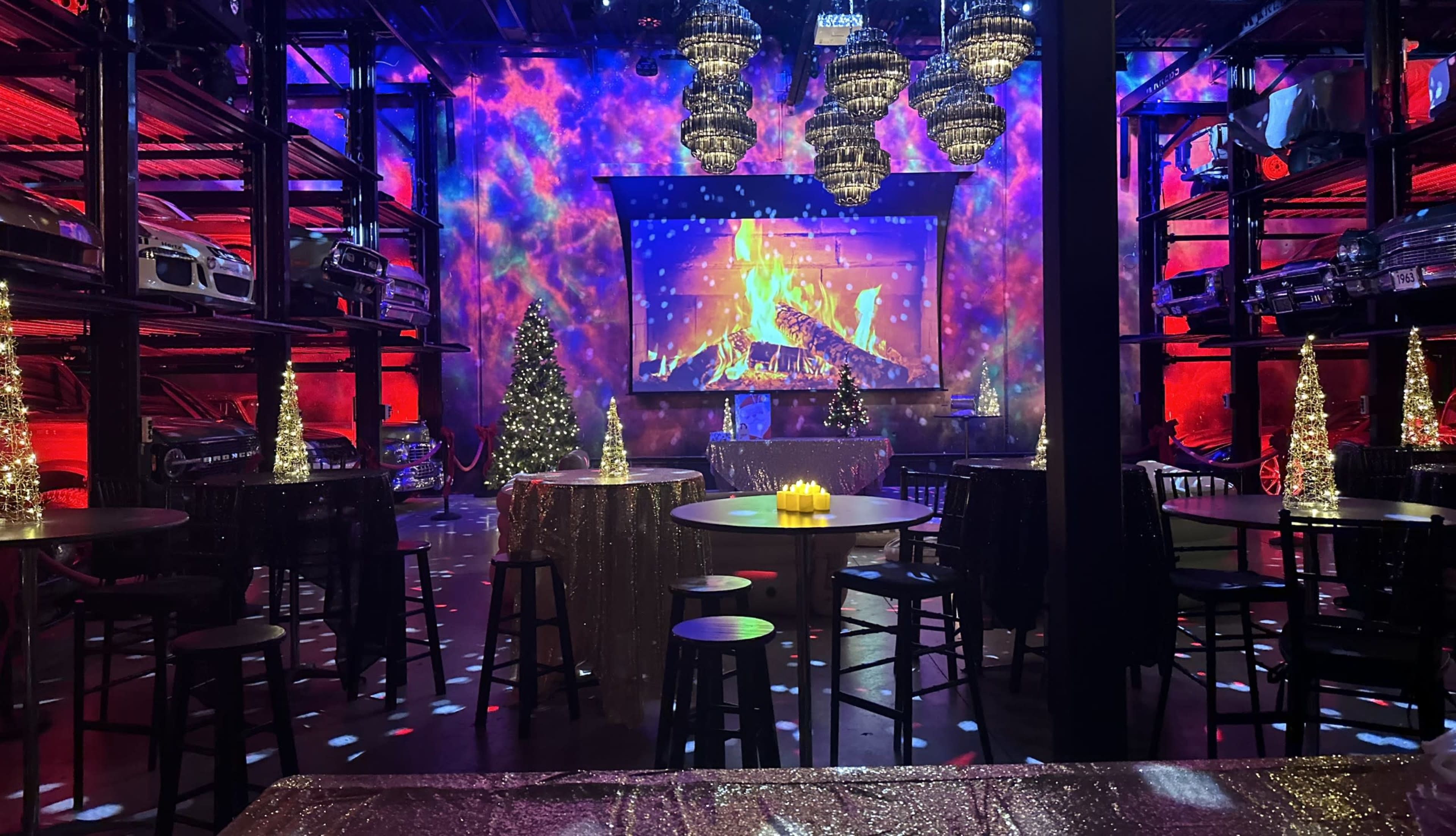 A festive interior of a venue decorated with colorful lights, a large screen displaying a fireplace scene, and tables with shimmering tablecloths surrounded by Christmas trees and sparkling chandeliers.