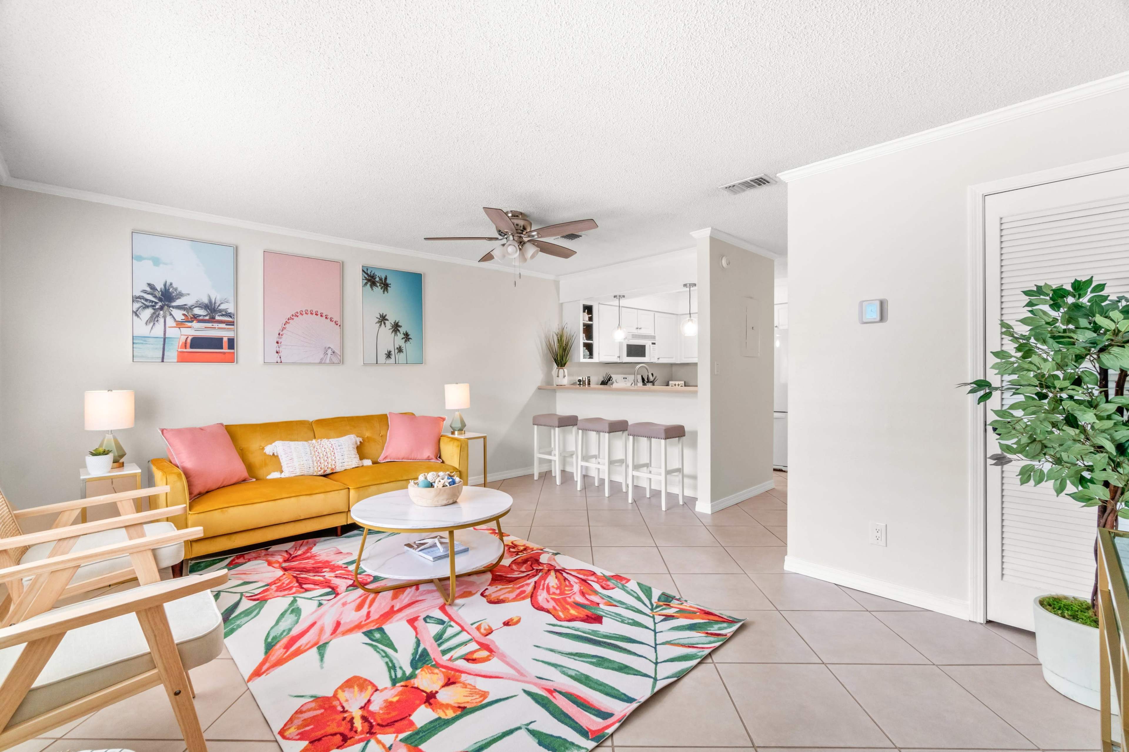 A bright living room features a yellow sofa, a floral rug, and a view into a kitchen with bar seating.