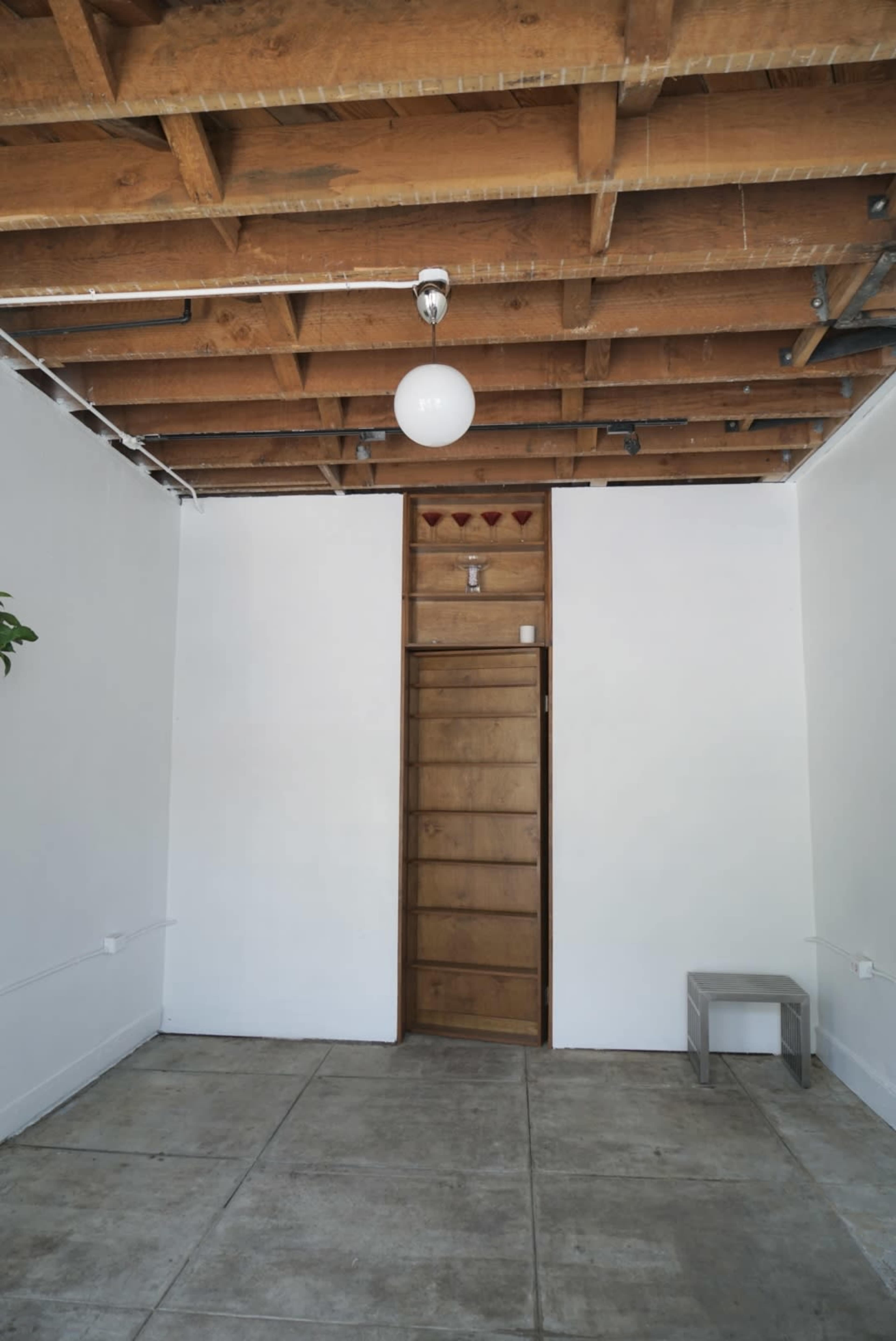 A minimalistic room with a wooden door, a simple light fixture, and a small bench against a white wall.