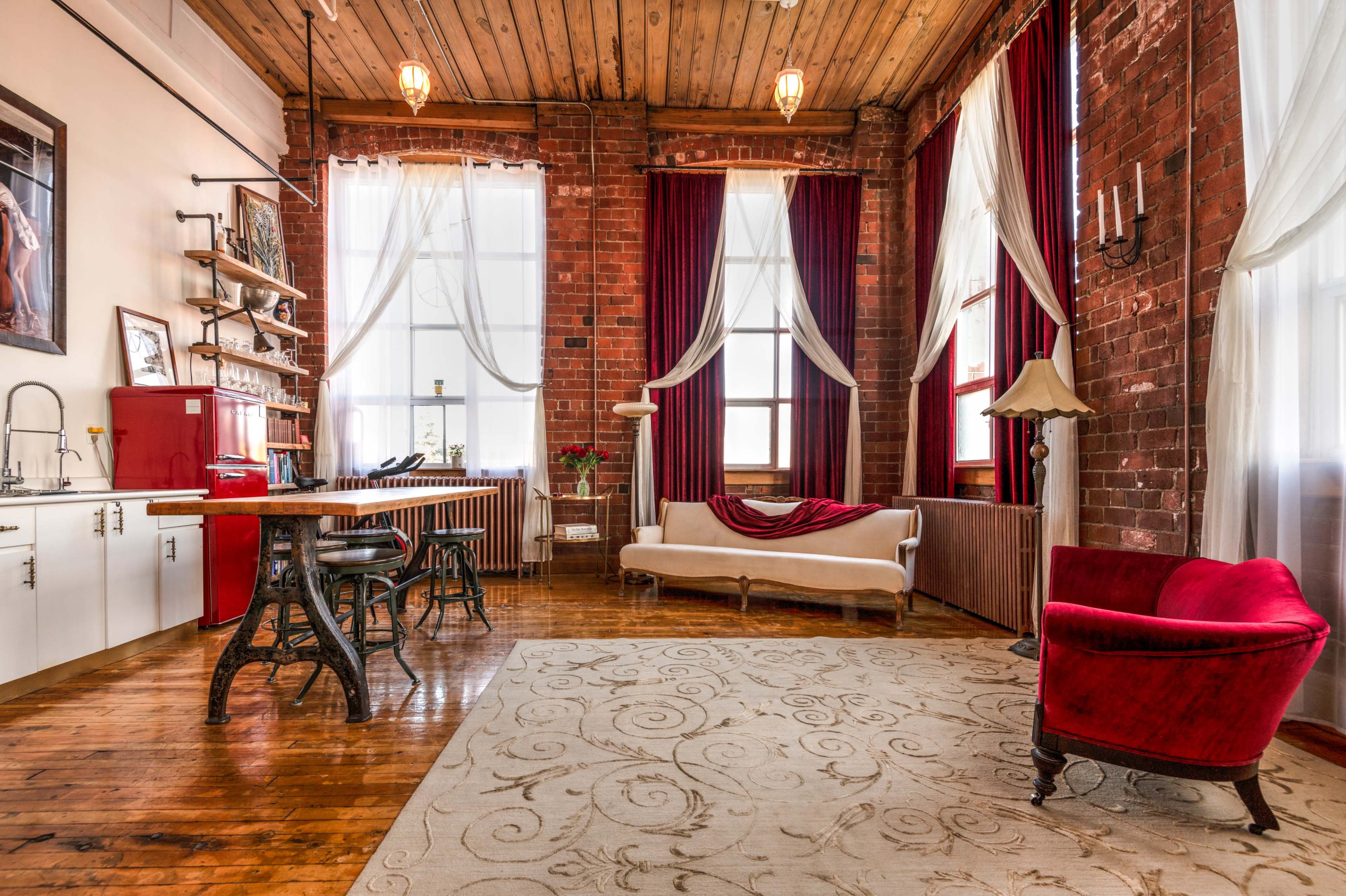 A spacious, industrial-style living area with exposed brick walls, wooden beams, and large windows, furnished with a white sofa, a red armchair, and a wooden dining table.