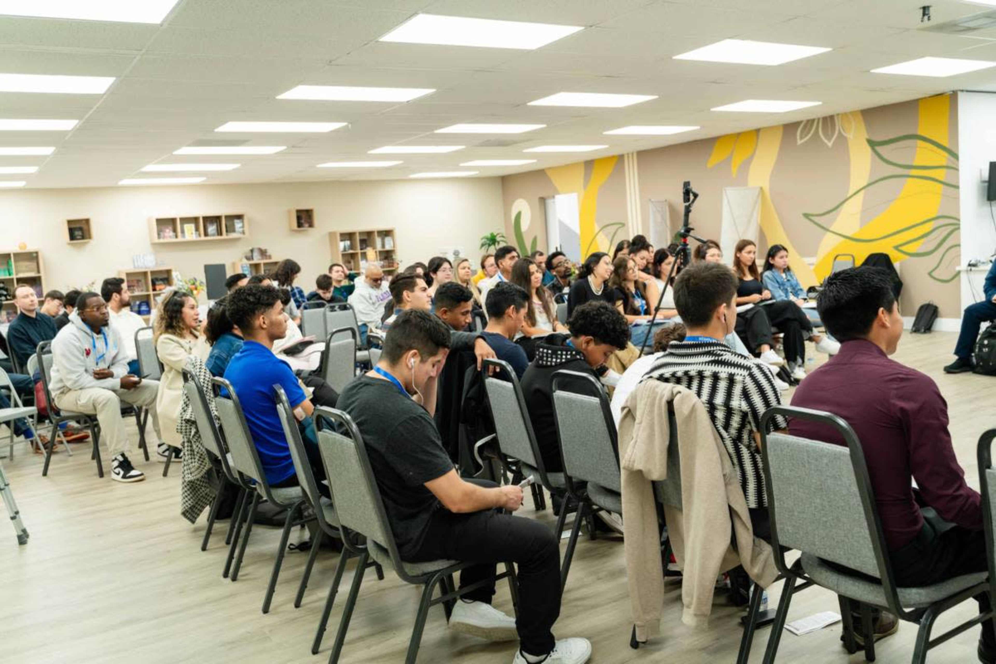 A group of people sits in rows during an event in a well-lit room with a mural on the wall.