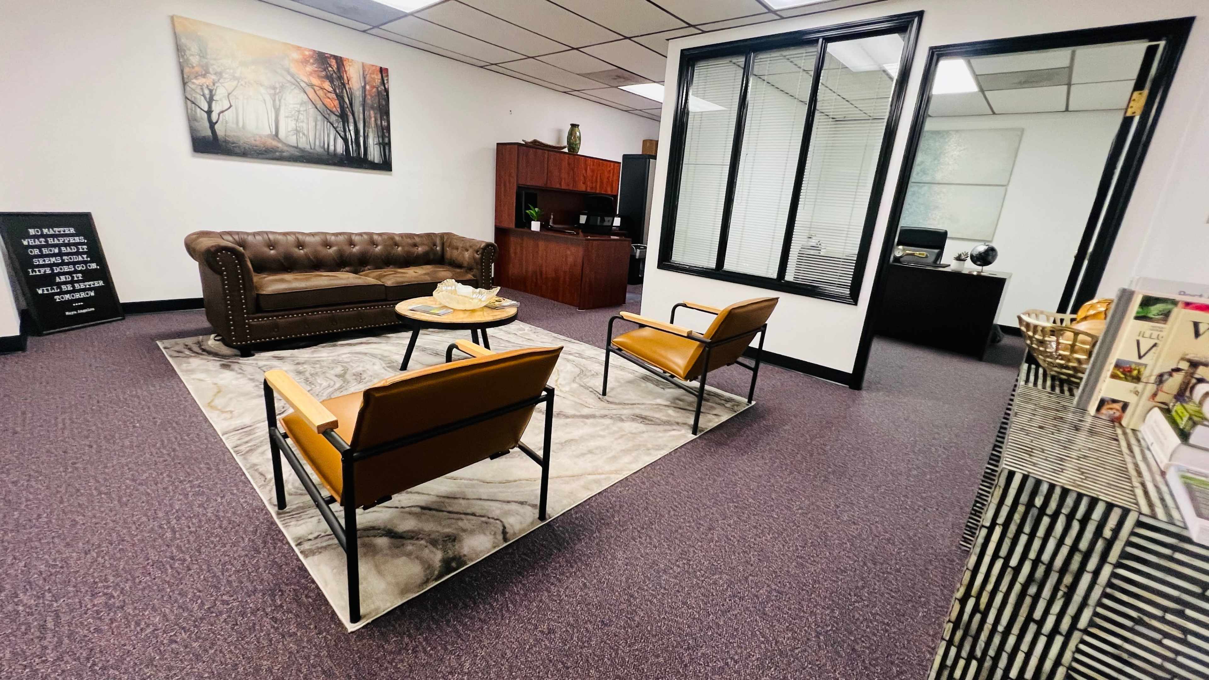 A professional office waiting area with a brown leather sofa, two orange chairs, a coffee table on a patterned rug, and a glass-walled office in the background.