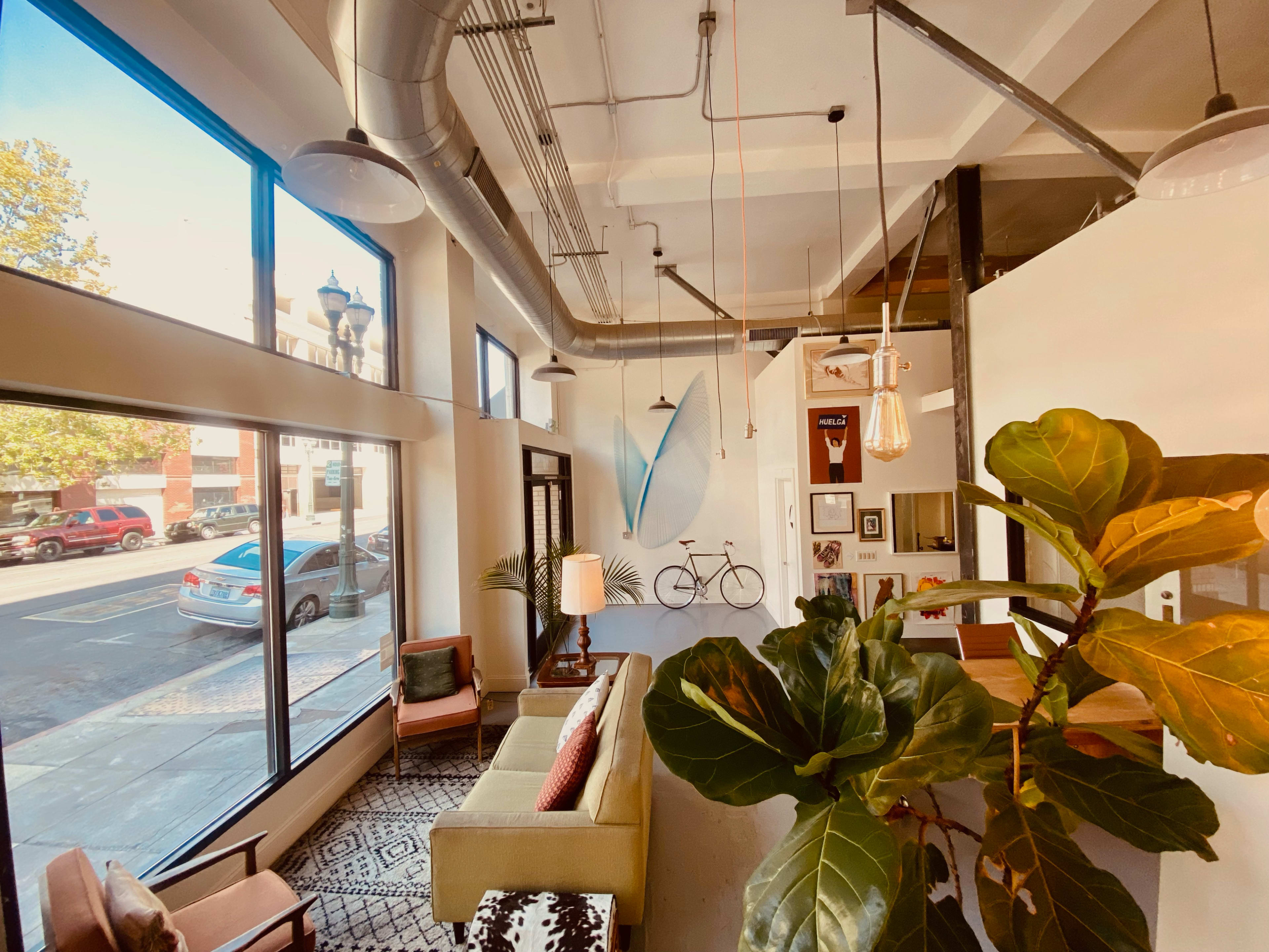 A bright, modern interior of a mixed-use space featuring a green sofa, a bicycle mounted on the wall, and large windows with views of the street.