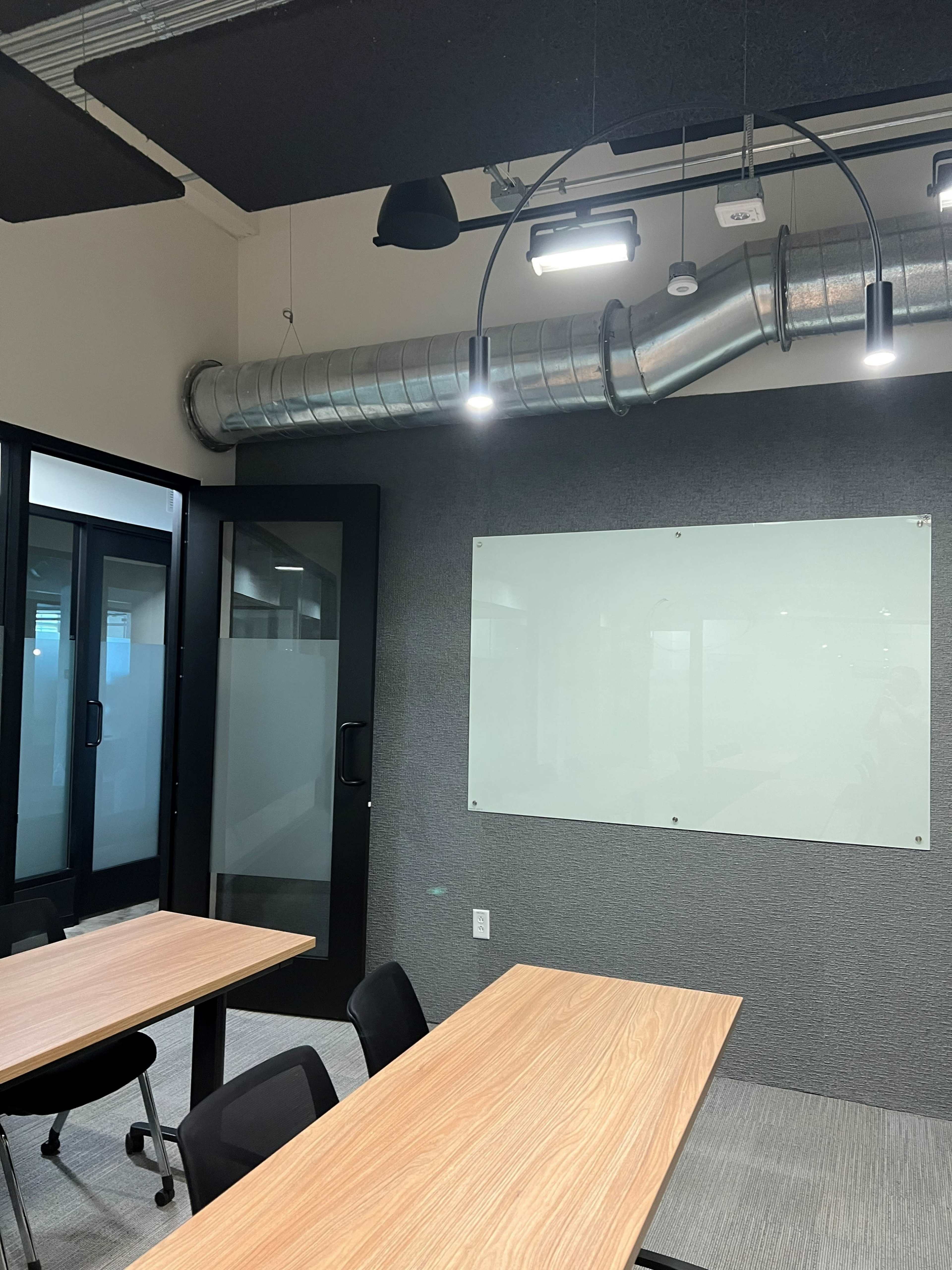 The image shows a modern conference room with two wooden tables, black chairs, and a glass whiteboard on a gray wall.