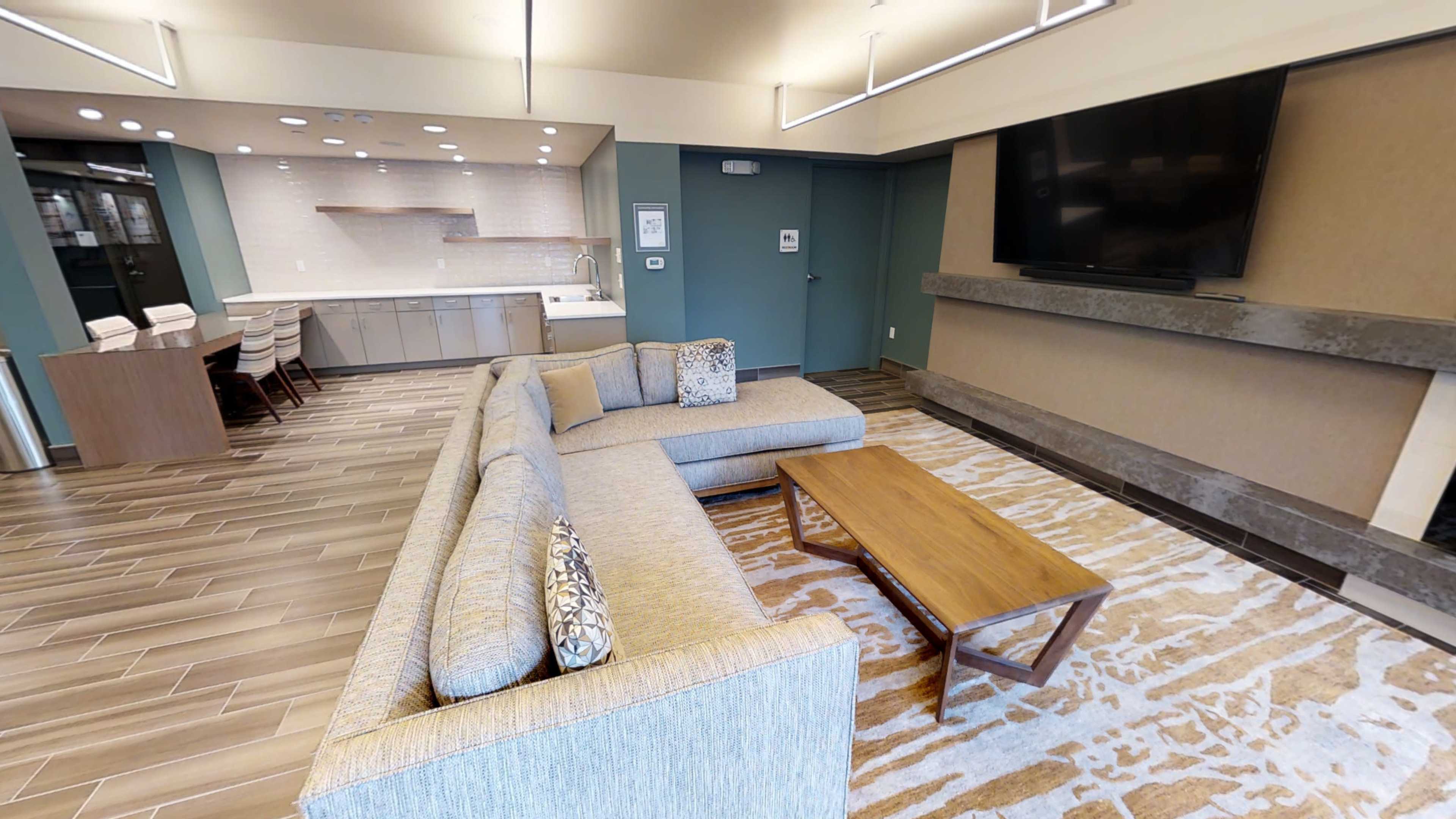 The image shows a modern lounge area with a sectional sofa, a wooden coffee table, and a wall-mounted television, adjacent to a kitchen space with bar seating.