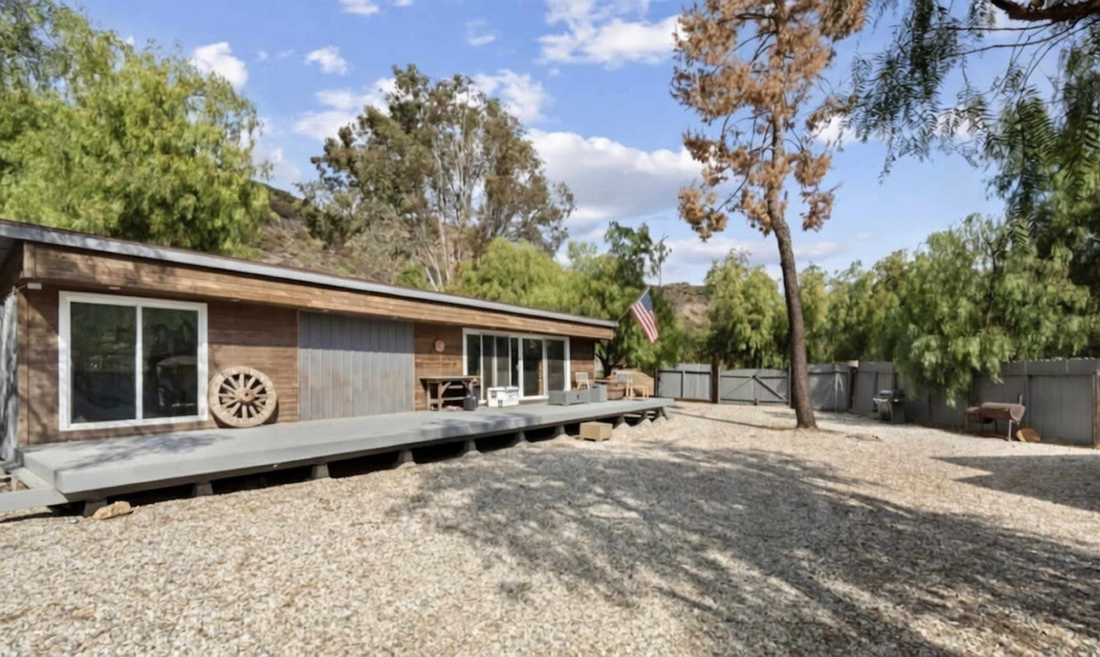 A wooden house with a large deck and an American flag is surrounded by gravel and trees in a rural setting.