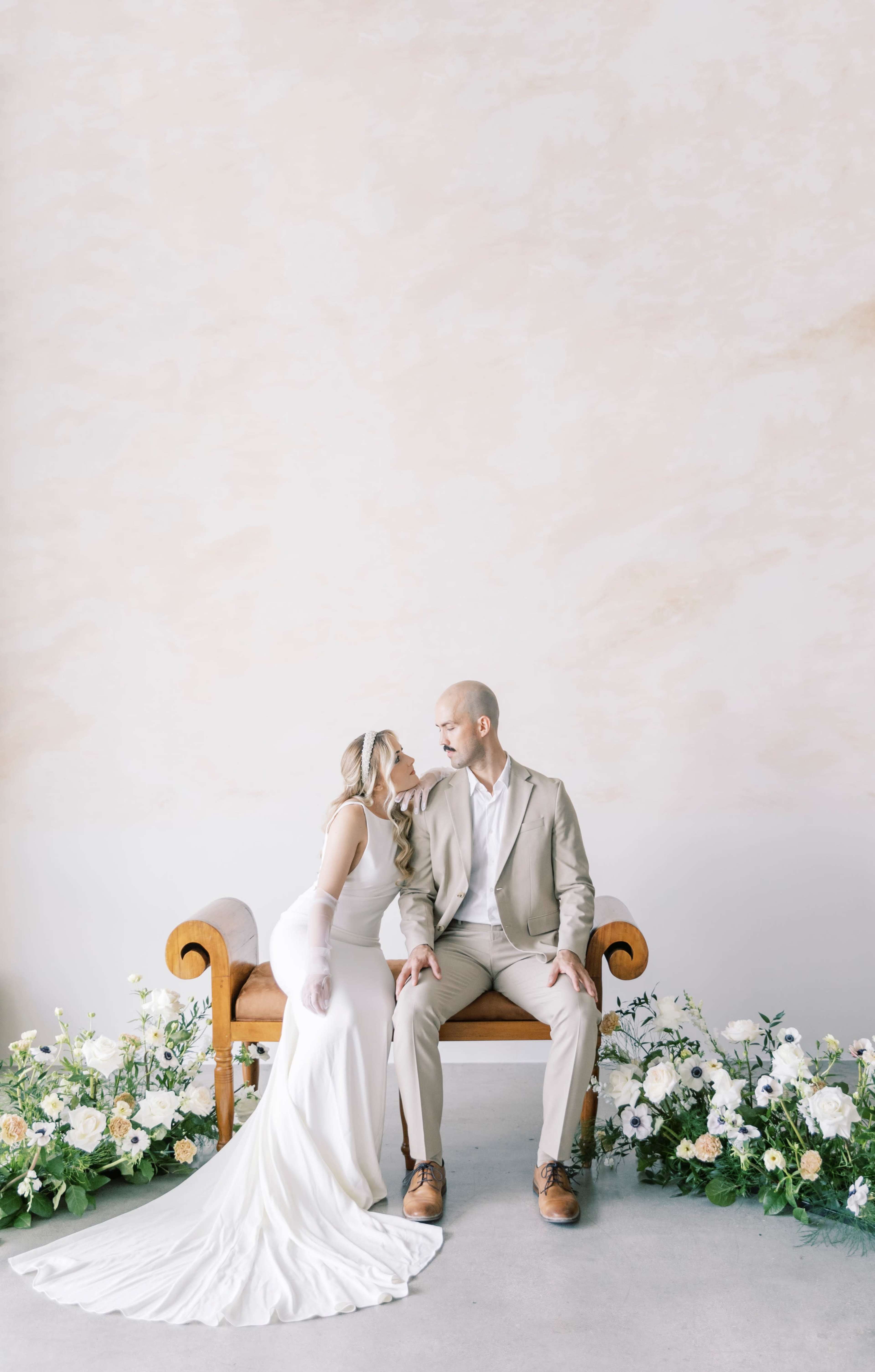 A couple sits together on a wooden bench surrounded by floral arrangements.